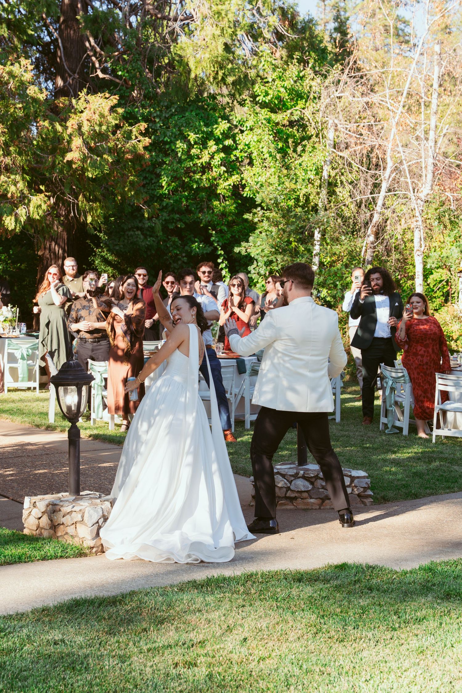 Wedding party celebrates outdoors in a park setting with a white-gowned bride and guests gathered on a path.