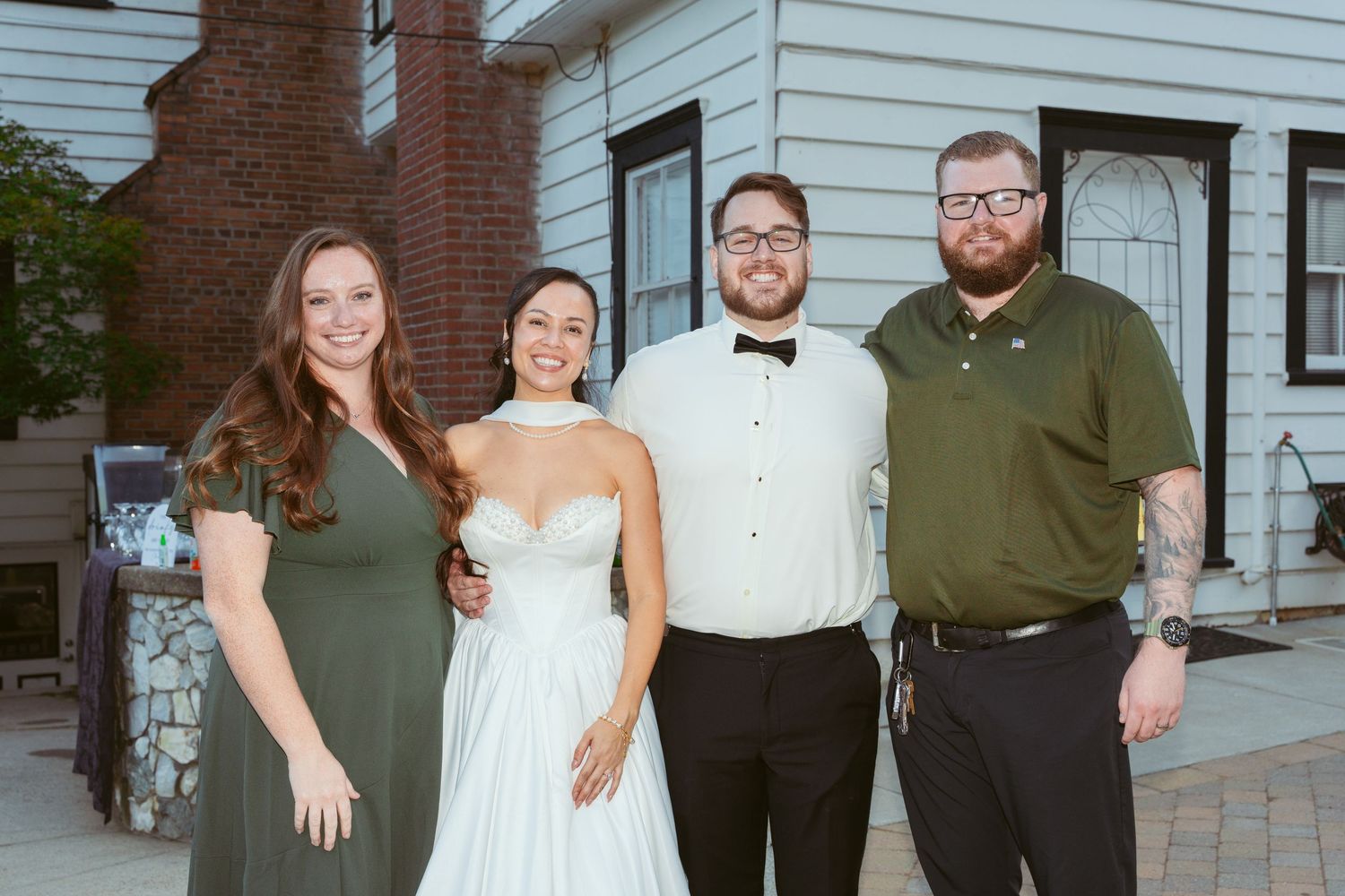 Wedding party members pose together outside a white house with brick accents.