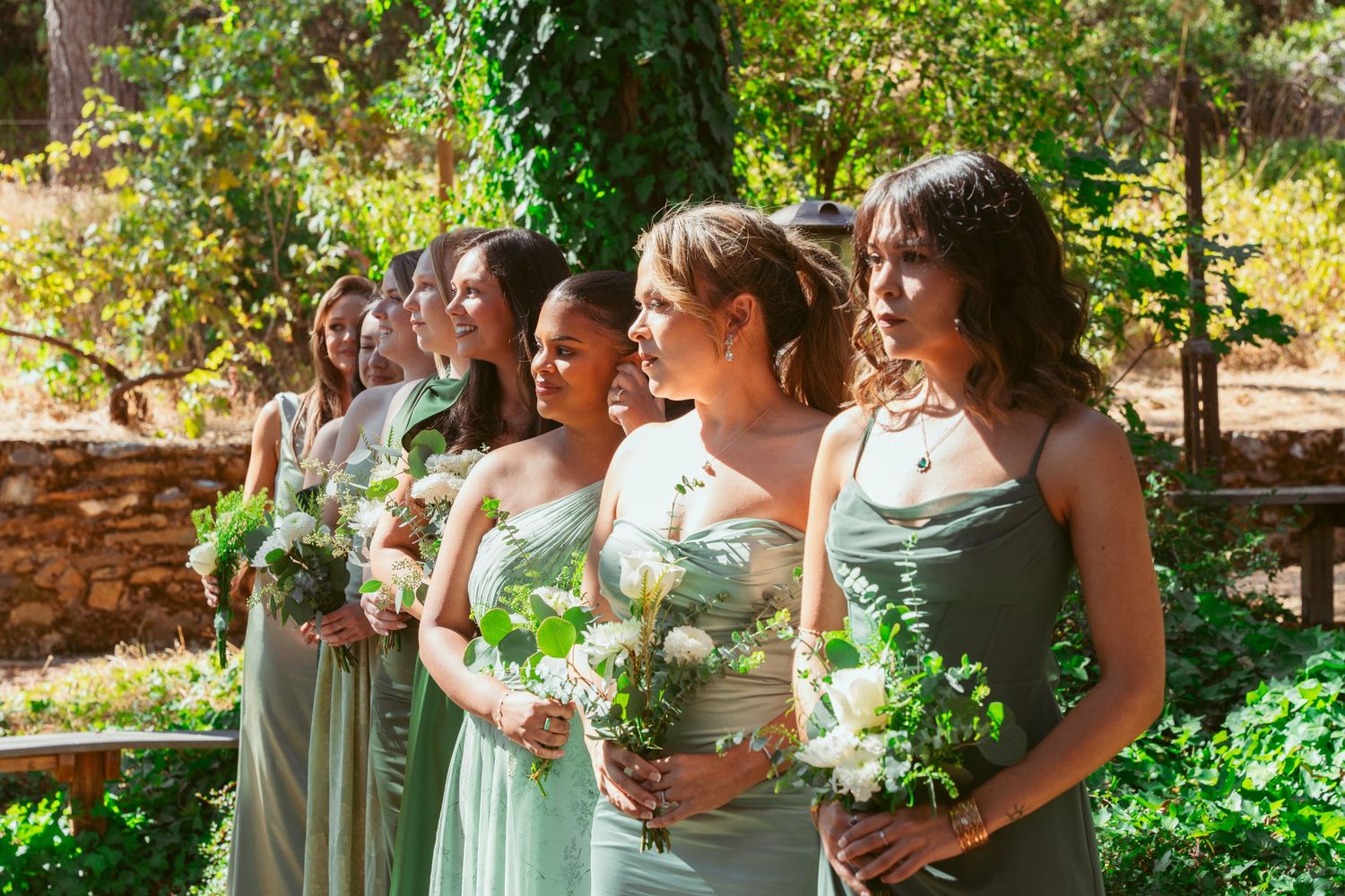 Bridesmaids in mint green dresses holding white bouquets stand together in a garden setting.