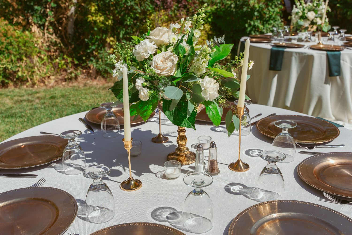 Elegant outdoor table setting with brown plates, wine glasses, and white floral centerpiece in brass candlesticks.
