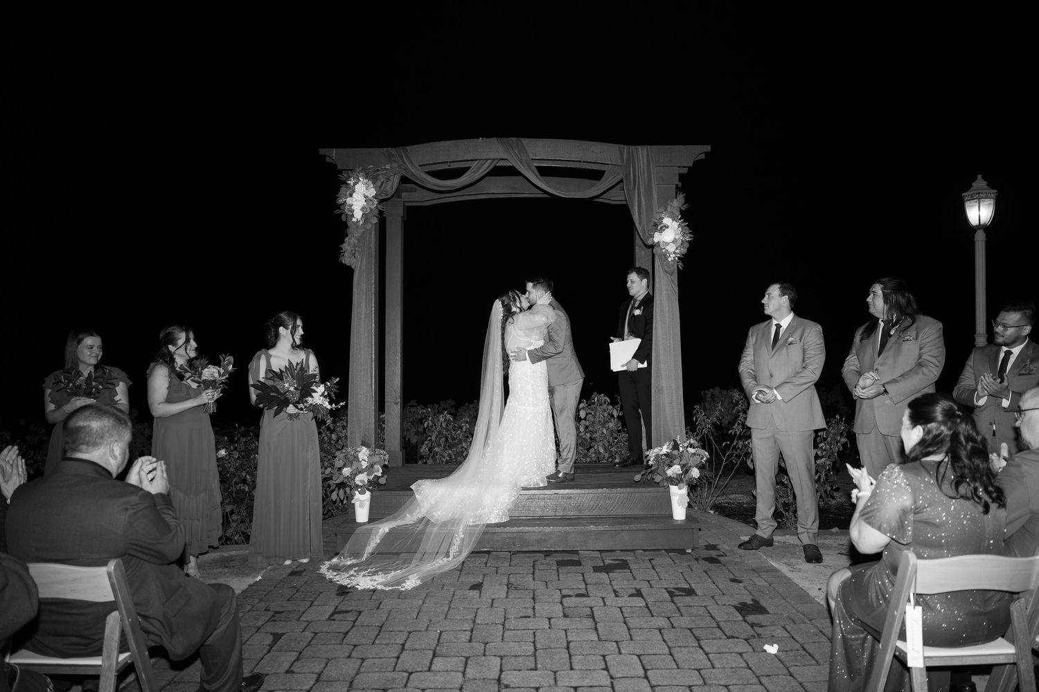 Wedding ceremony taking place at dusk with guests watching from chairs on brick patio.