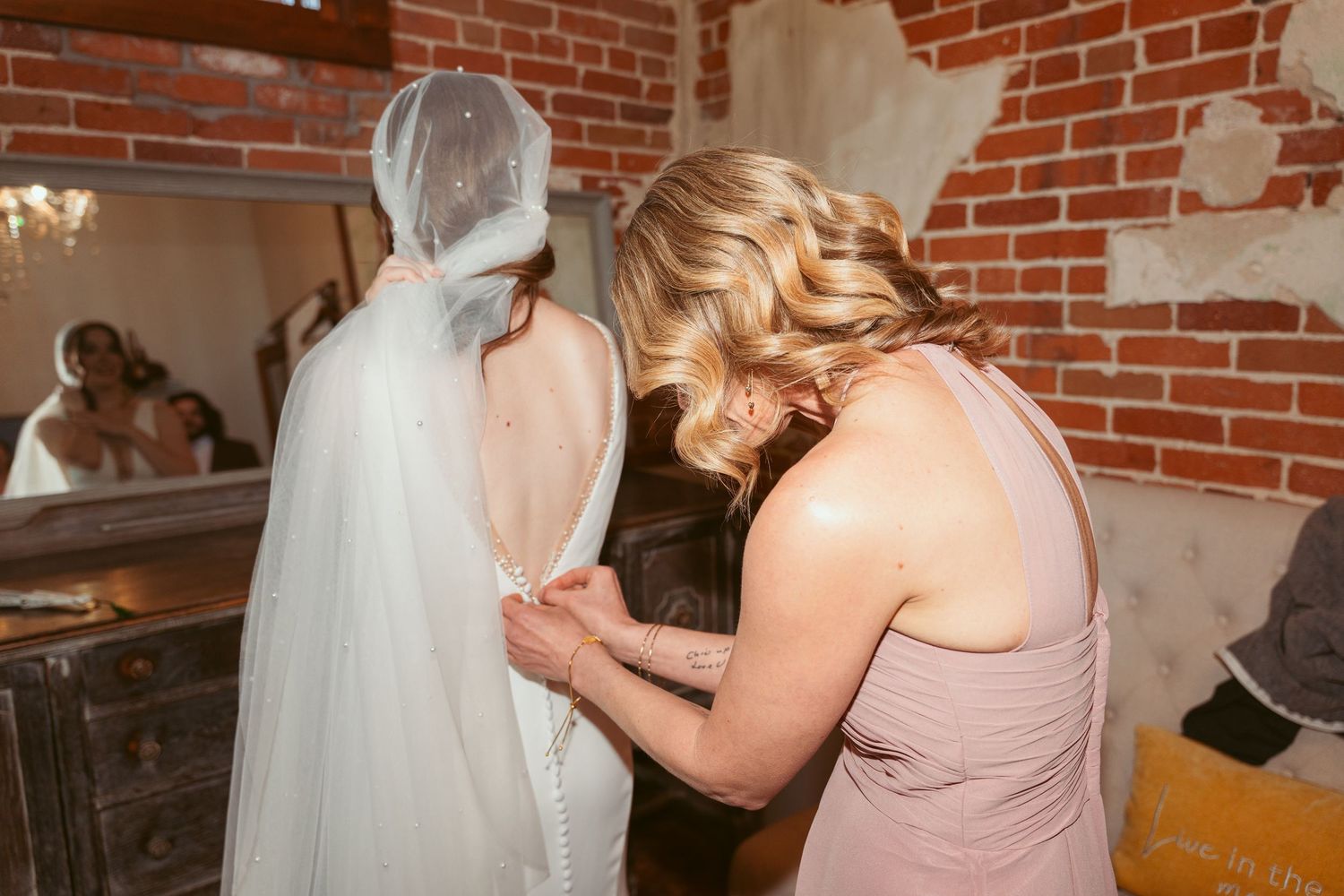 Wedding party members helping with dress preparations against exposed brick wall.