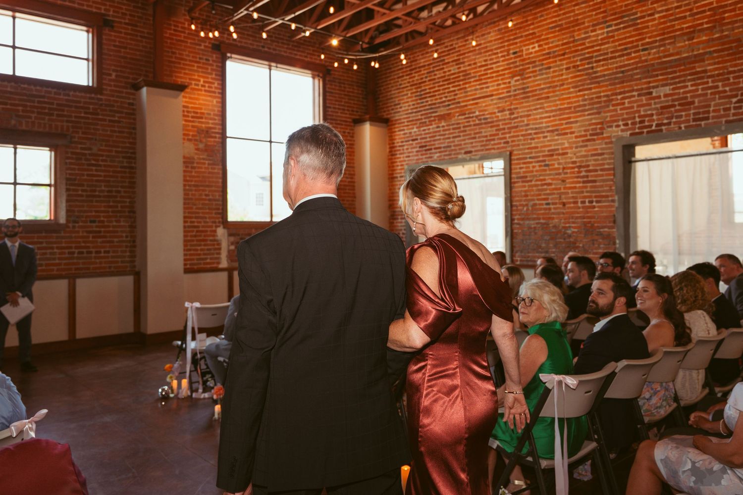 Indoor wedding ceremony in a rustic brick venue with string lights overhead.