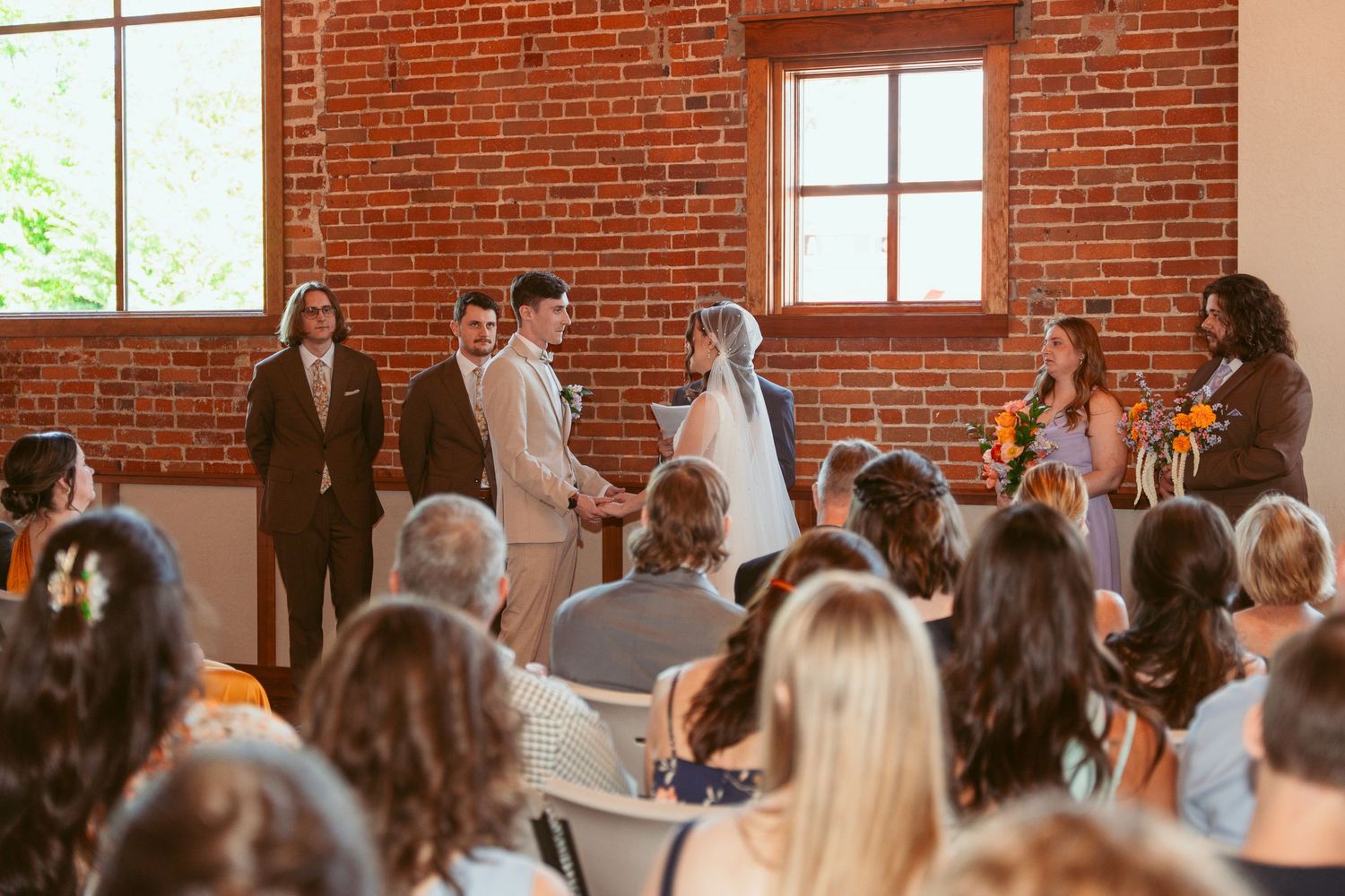 Wedding guests observing ceremony in historic venue with exposed brick walls and tall windows.