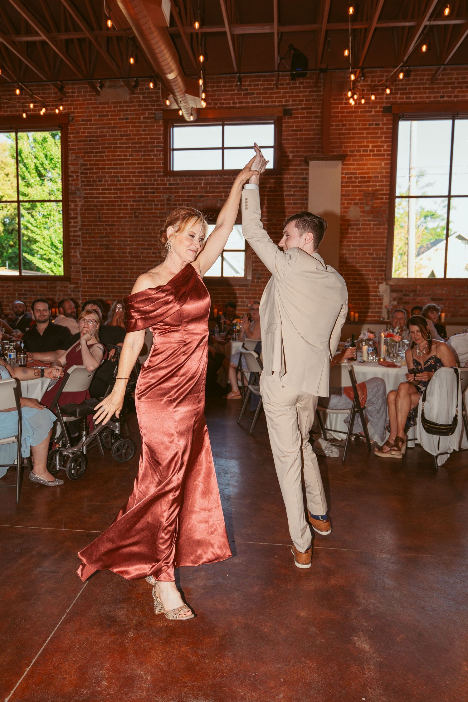 Wedding guests dancing energetically on a dark wooden floor in a rustic brick venue with string lights.