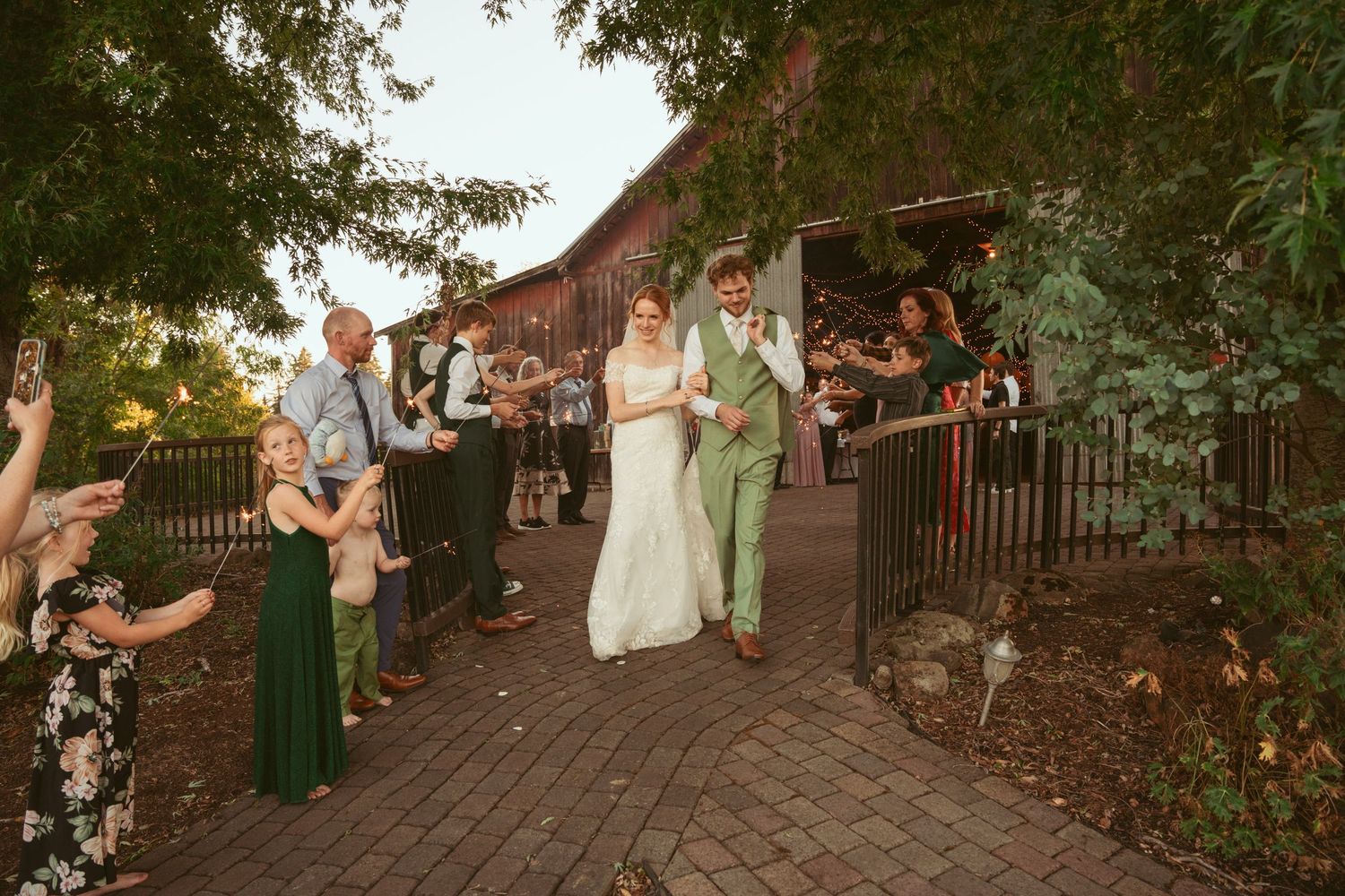 Wedding guests line pathway with sparklers as couple exits rustic barn venue at sunset.