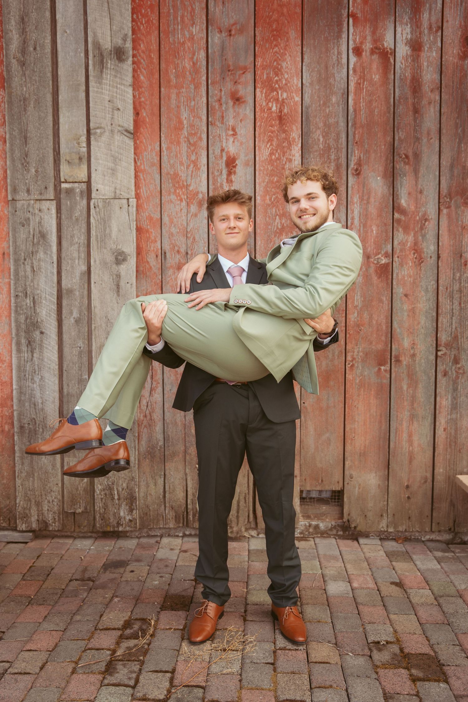 Playful wedding portrait with one person carrying another in front of barn wall.