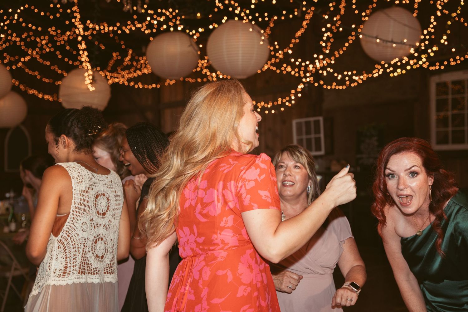 Wedding guests dancing and celebrating under warm string lights at an indoor reception.