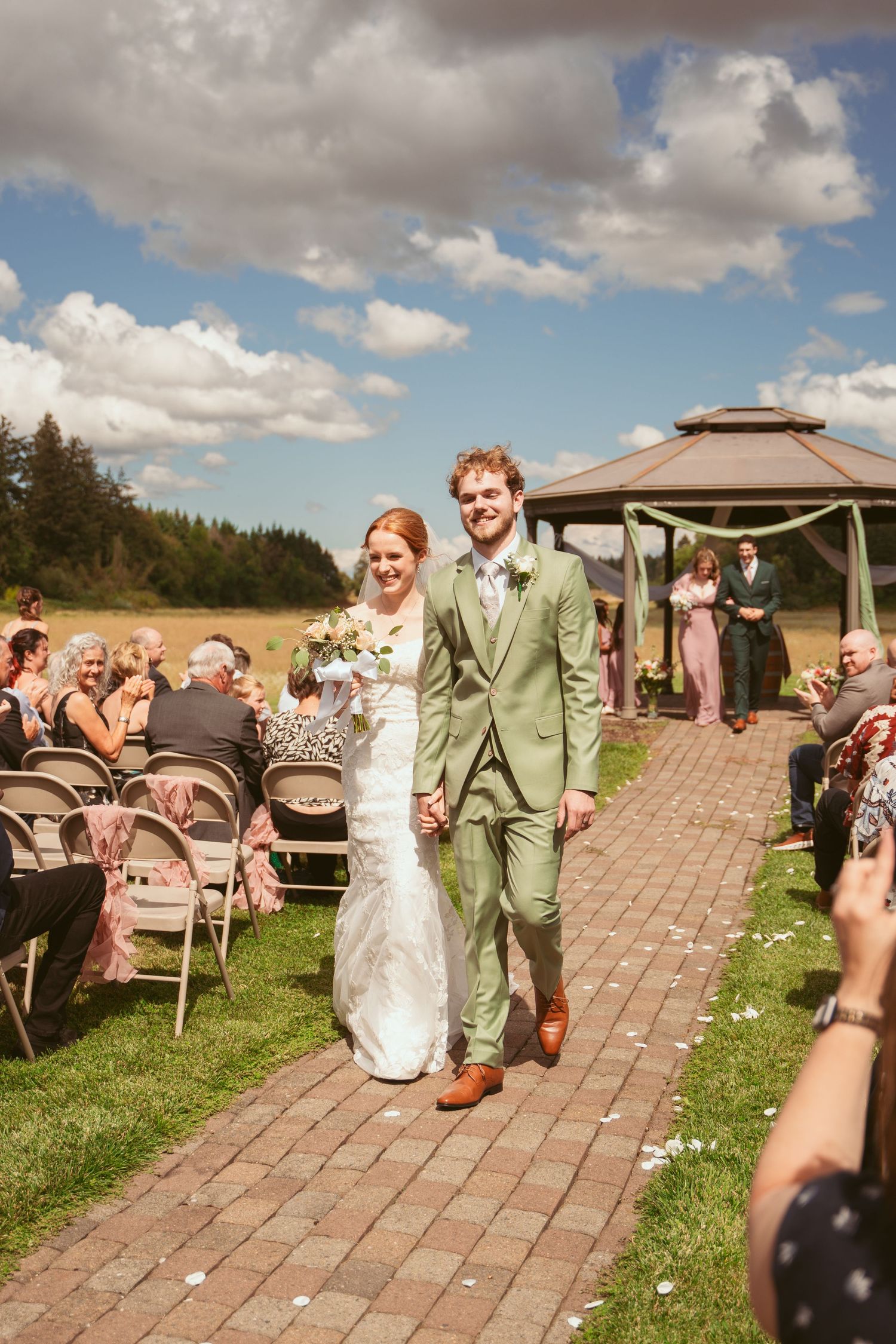 Newlyweds walking down brick aisle after outdoor gazebo ceremony with guests seated on both sides.