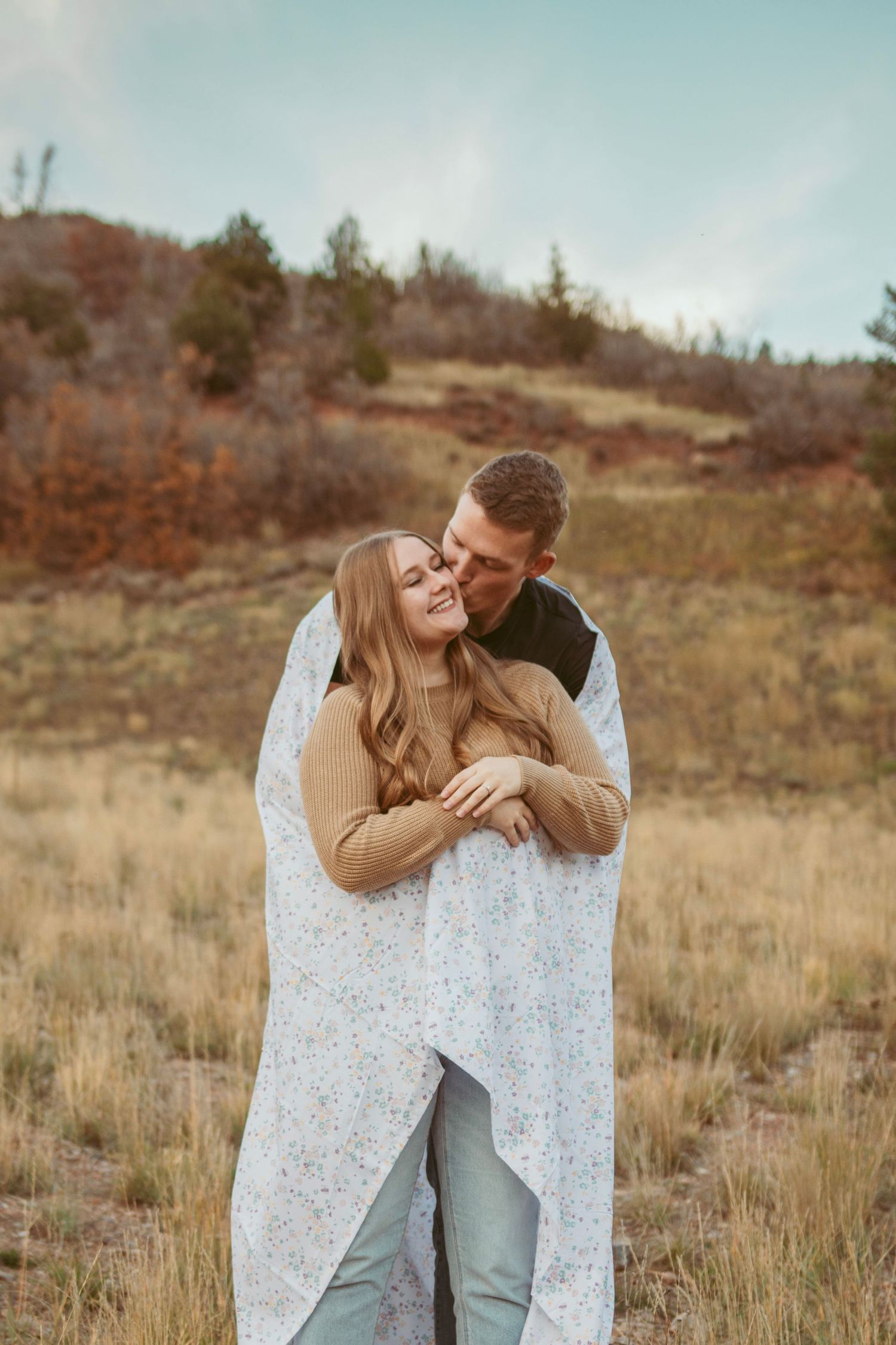 Playful couple embracing while draped in delicate white lace blanket against mountain backdrop.