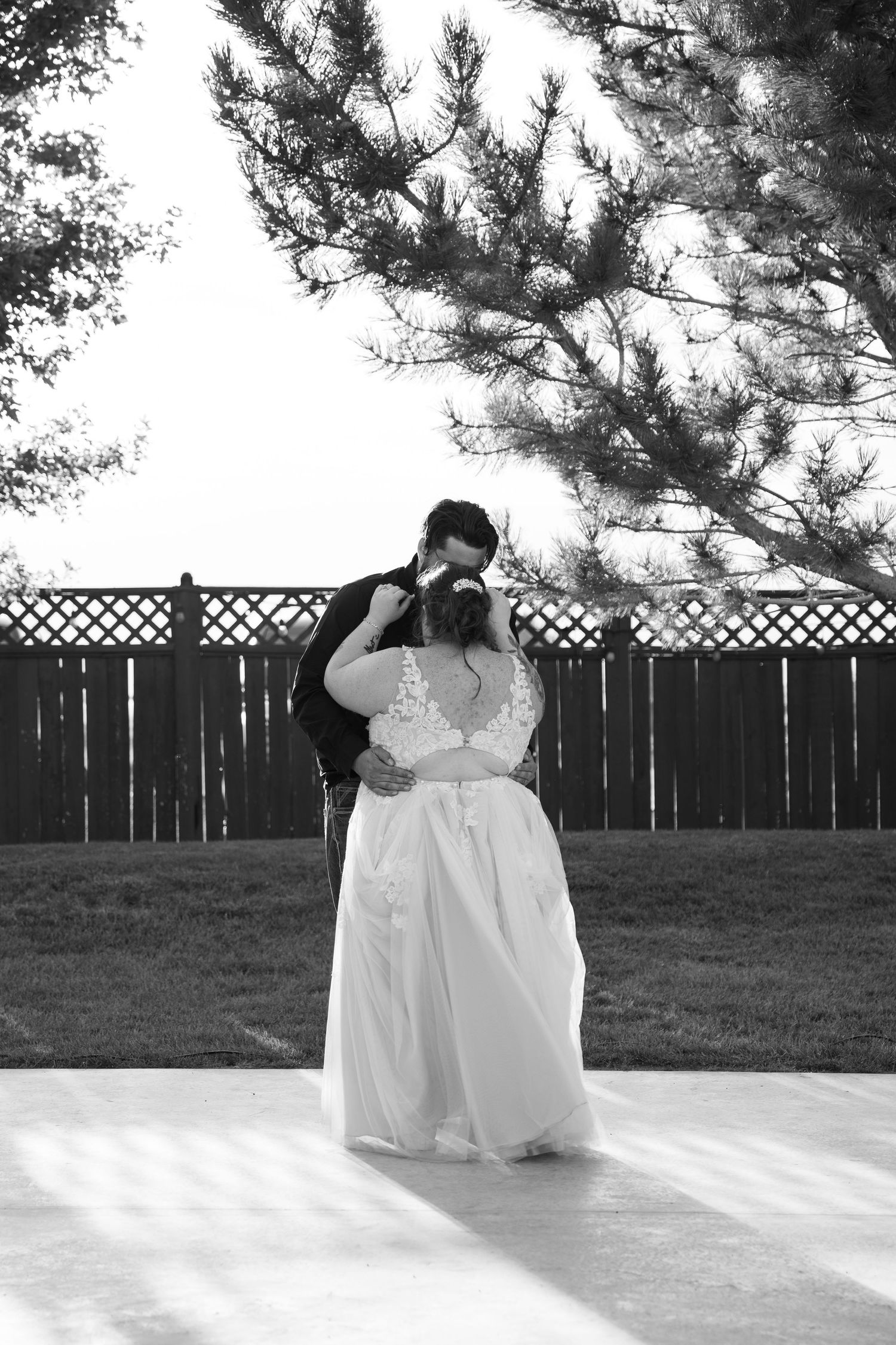 Black and white wedding photo with pine trees and lattice fence in background.