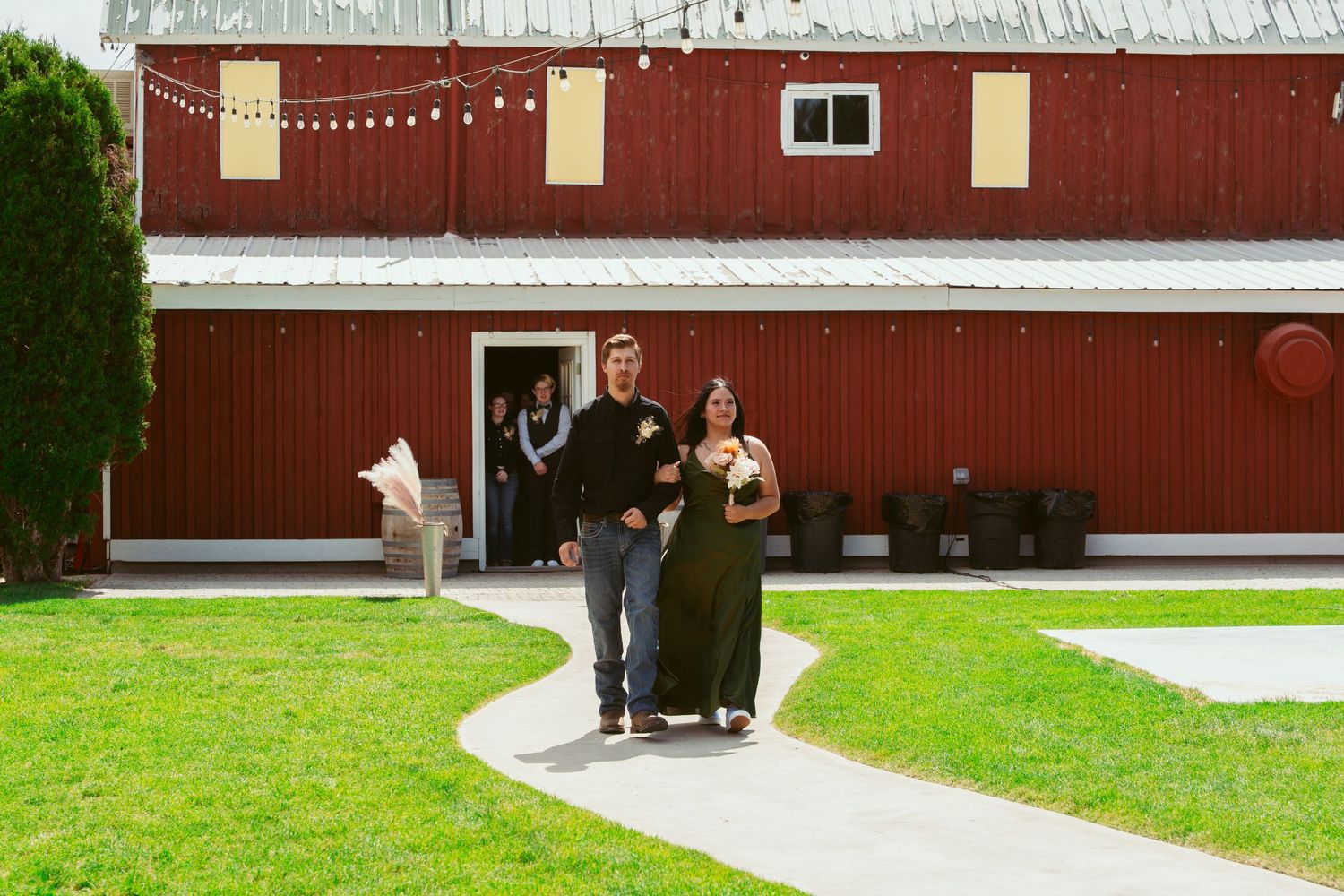 Couple exiting a red barn venue decorated with string lights during a wedding celebration.