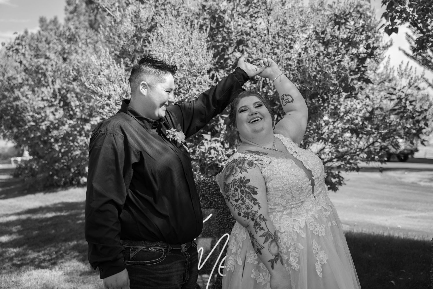 A joyful couple poses outdoors in a garden setting, with one wearing a black jacket and the other in a lace wedding dress.