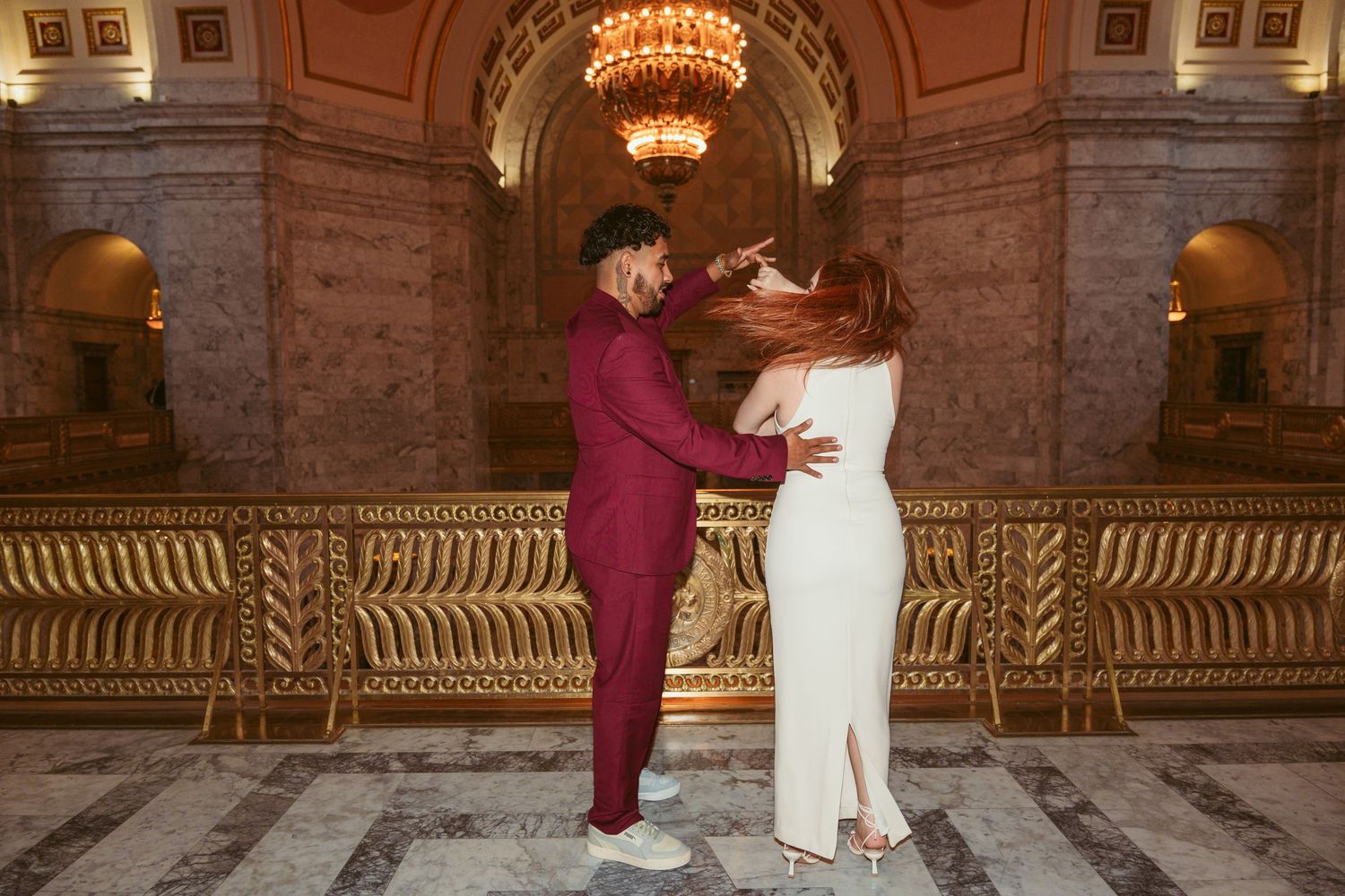 A couple in burgundy suit and white dress share an intimate moment under an ornate chandelier in a marble hallway.