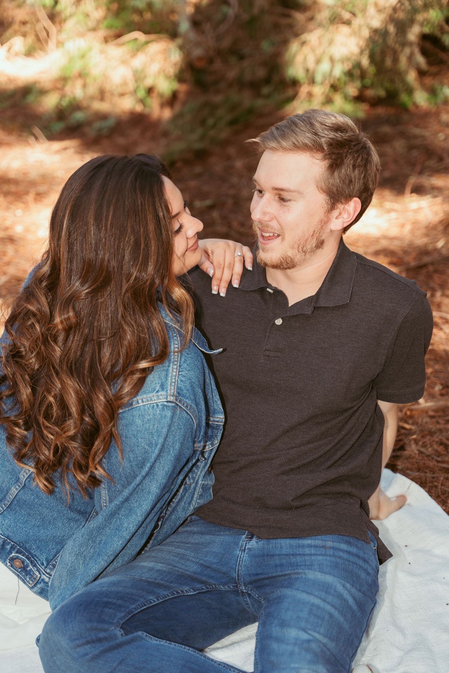 A couple in casual attire shares loving moments on a blanket in a wooded setting.