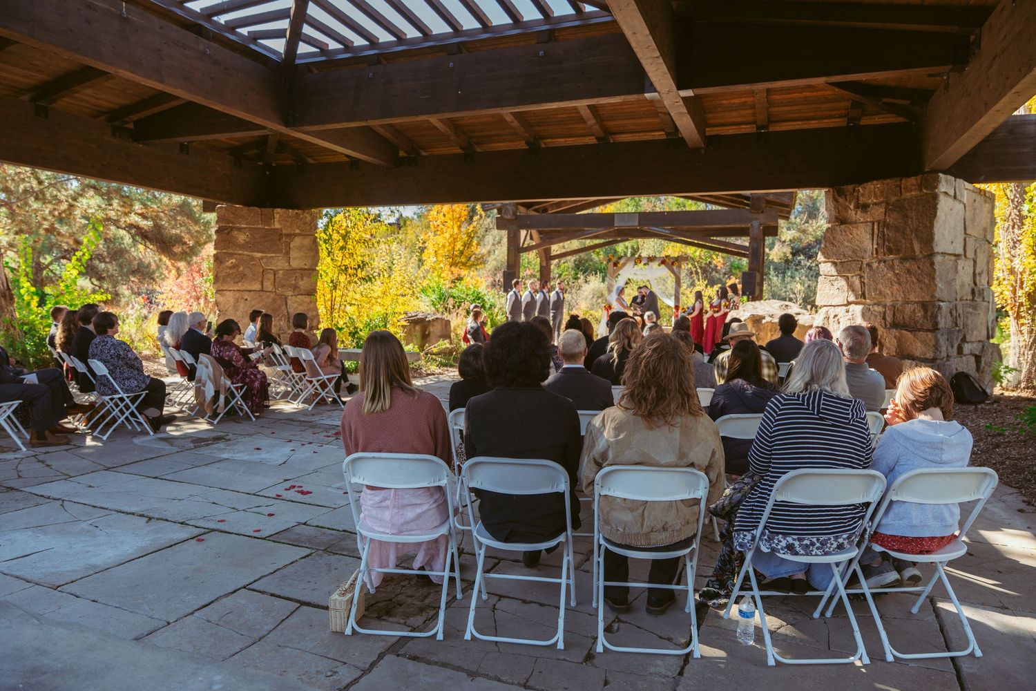 Side view of wedding guests seated in white chairs under covered stone pavilion.