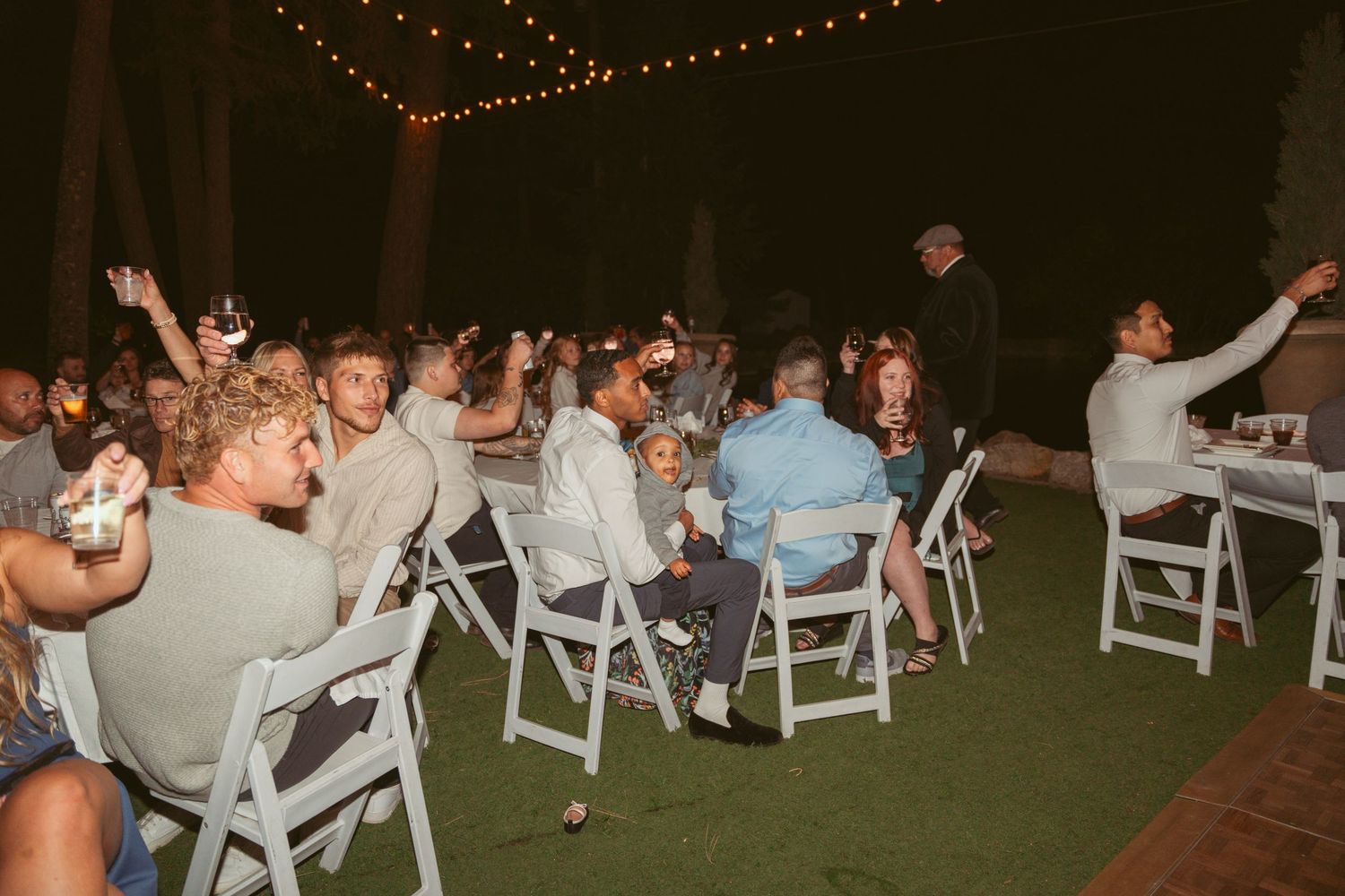 Wedding guests seated in white chairs at outdoor reception under string lights.
