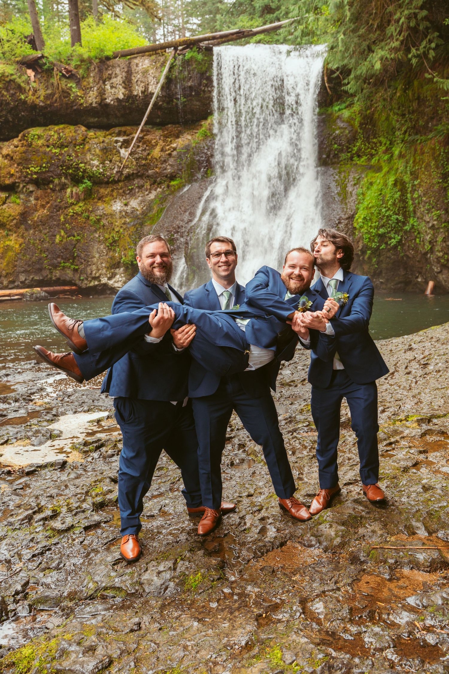 Wedding party members strike a fun pose by a waterfall in matching navy suits.
