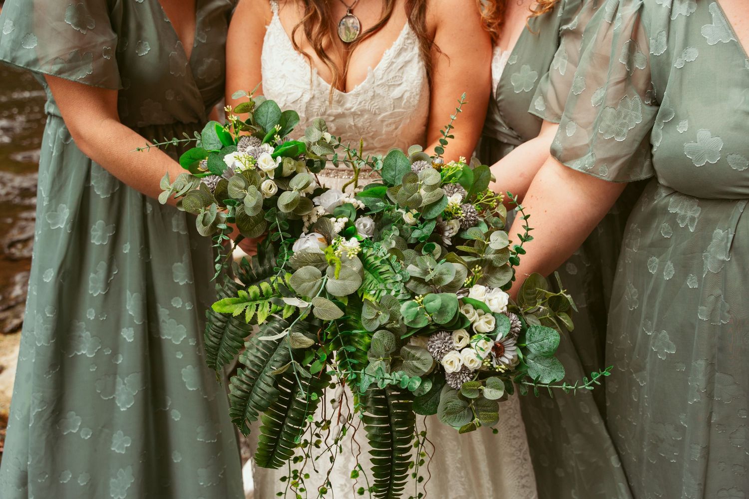 Wedding bouquets featuring lush greenery and white flowers held by people in sage green dresses.