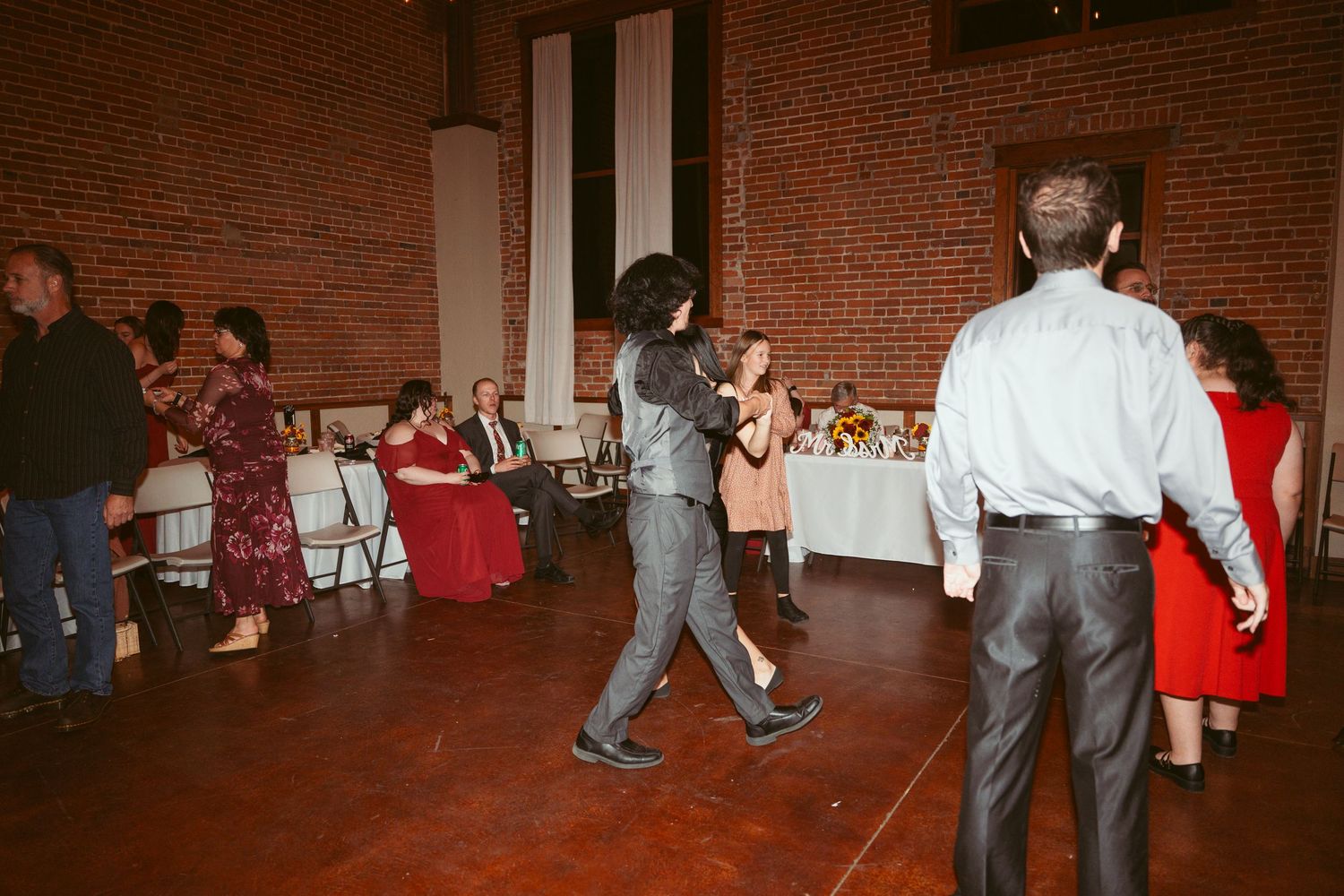 Couples dancing at a reception with exposed brick walls and white tablecloths in the background.