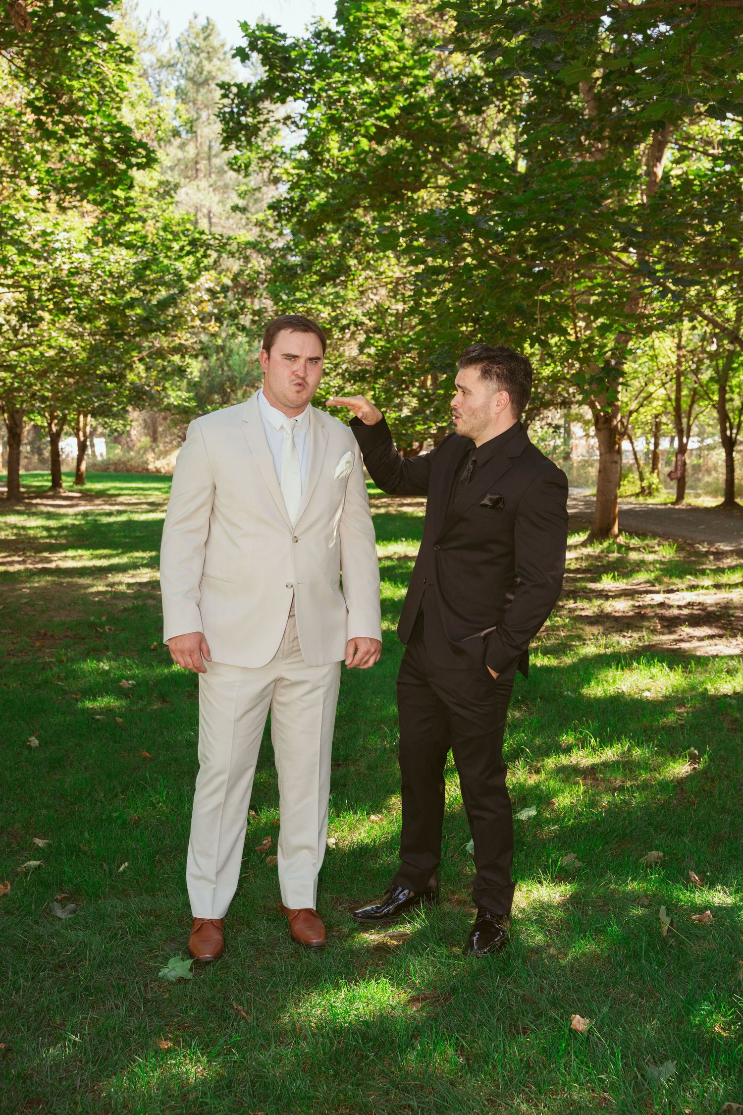 Two people in light and dark suits stand together on grass under leafy green trees.