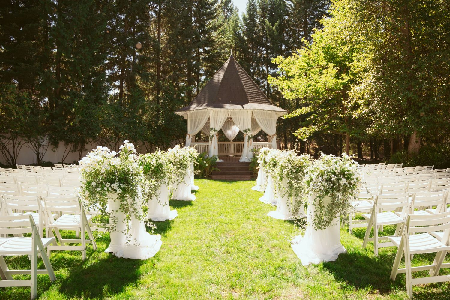Outdoor wedding ceremony setup with white chairs and hanging floral decorations in a sunny garden setting.