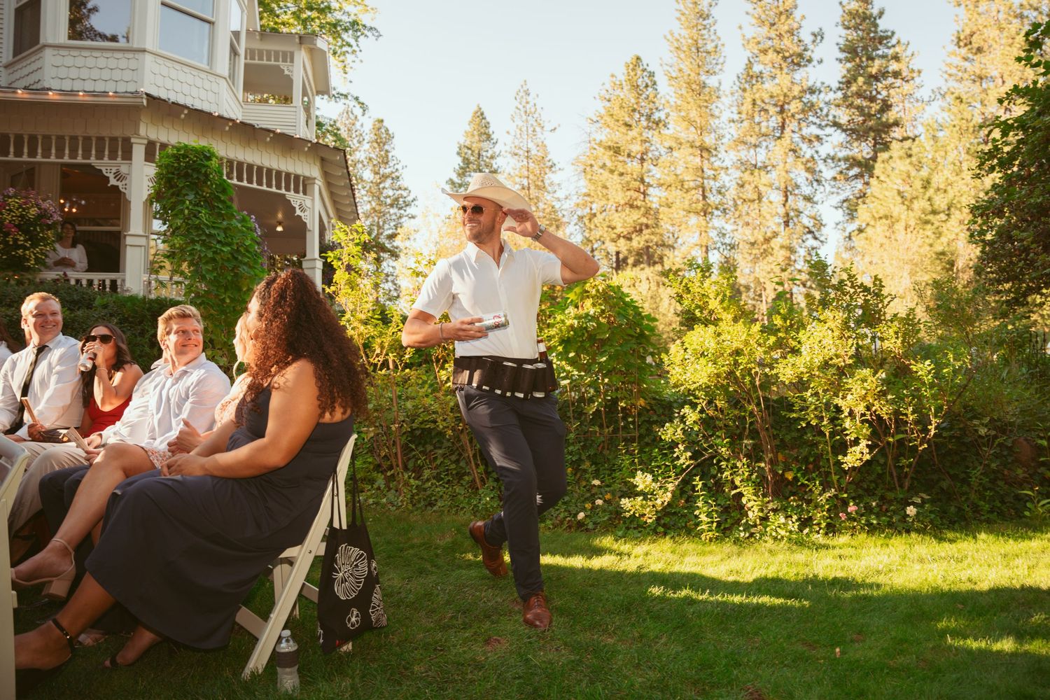 Wedding guests dance and celebrate outdoors at sunset with tall evergreen trees in the background.