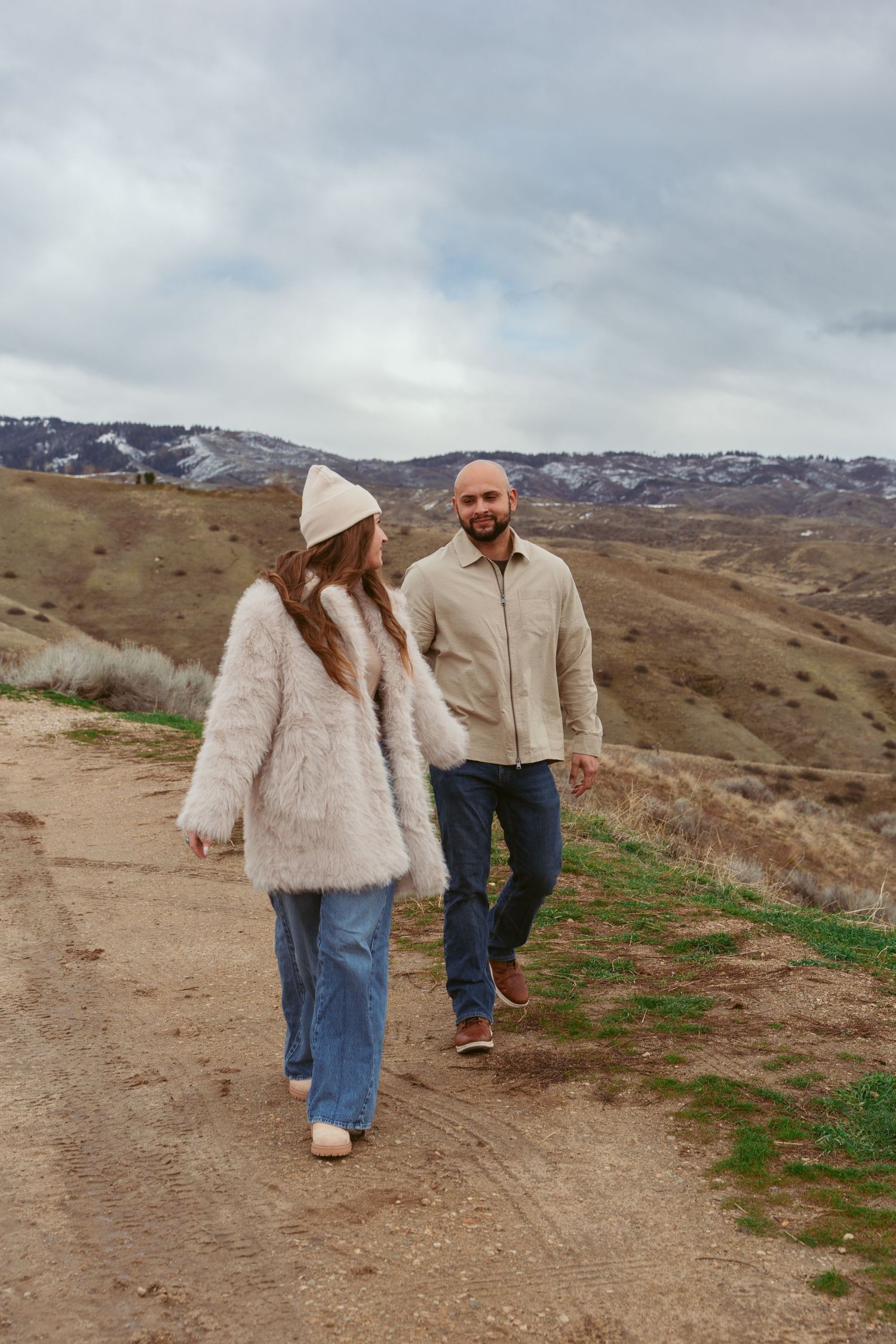A couple walks together on a mountain trail with snow-capped peaks in the background.