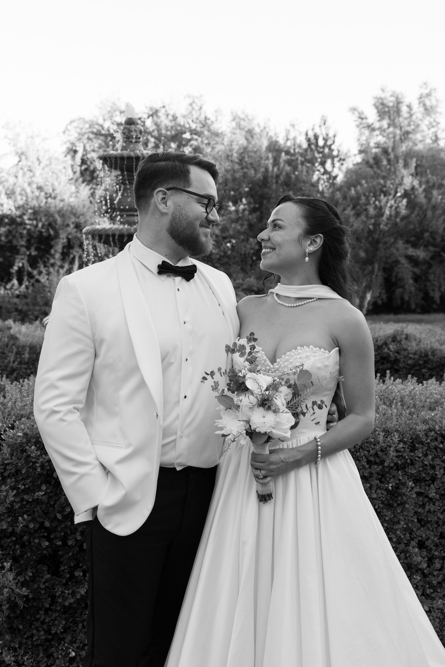 A black and white wedding portrait of a couple in formal attire sharing a loving moment in a garden setting.