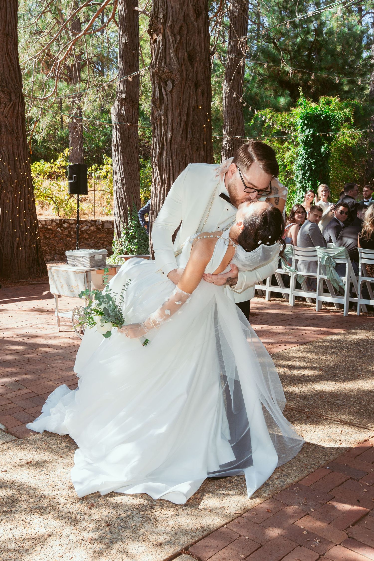 Romantic outdoor wedding photos taken under tall trees with white chairs and decorations.