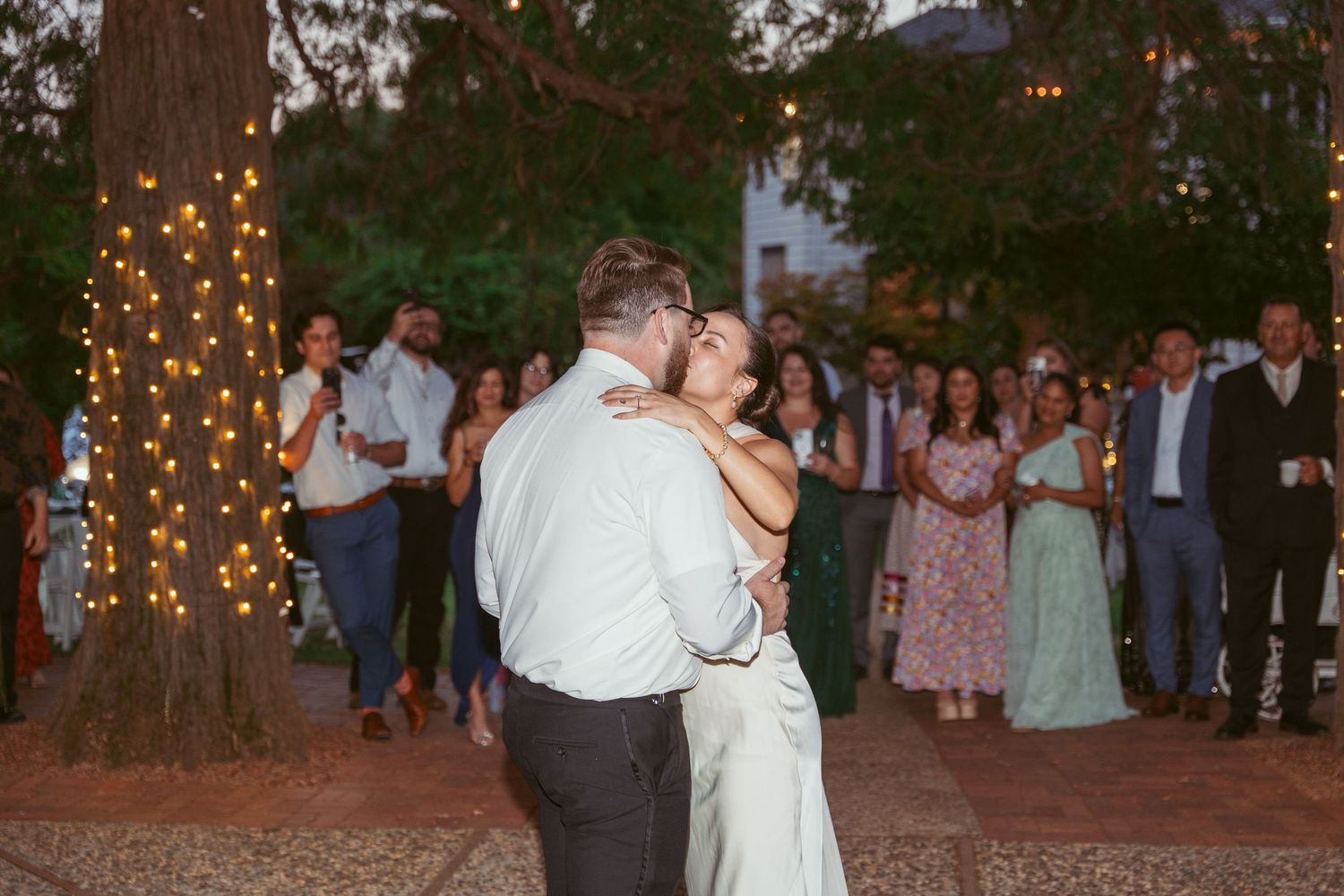 Couples slow dance at a romantic outdoor wedding reception under illuminated trees.