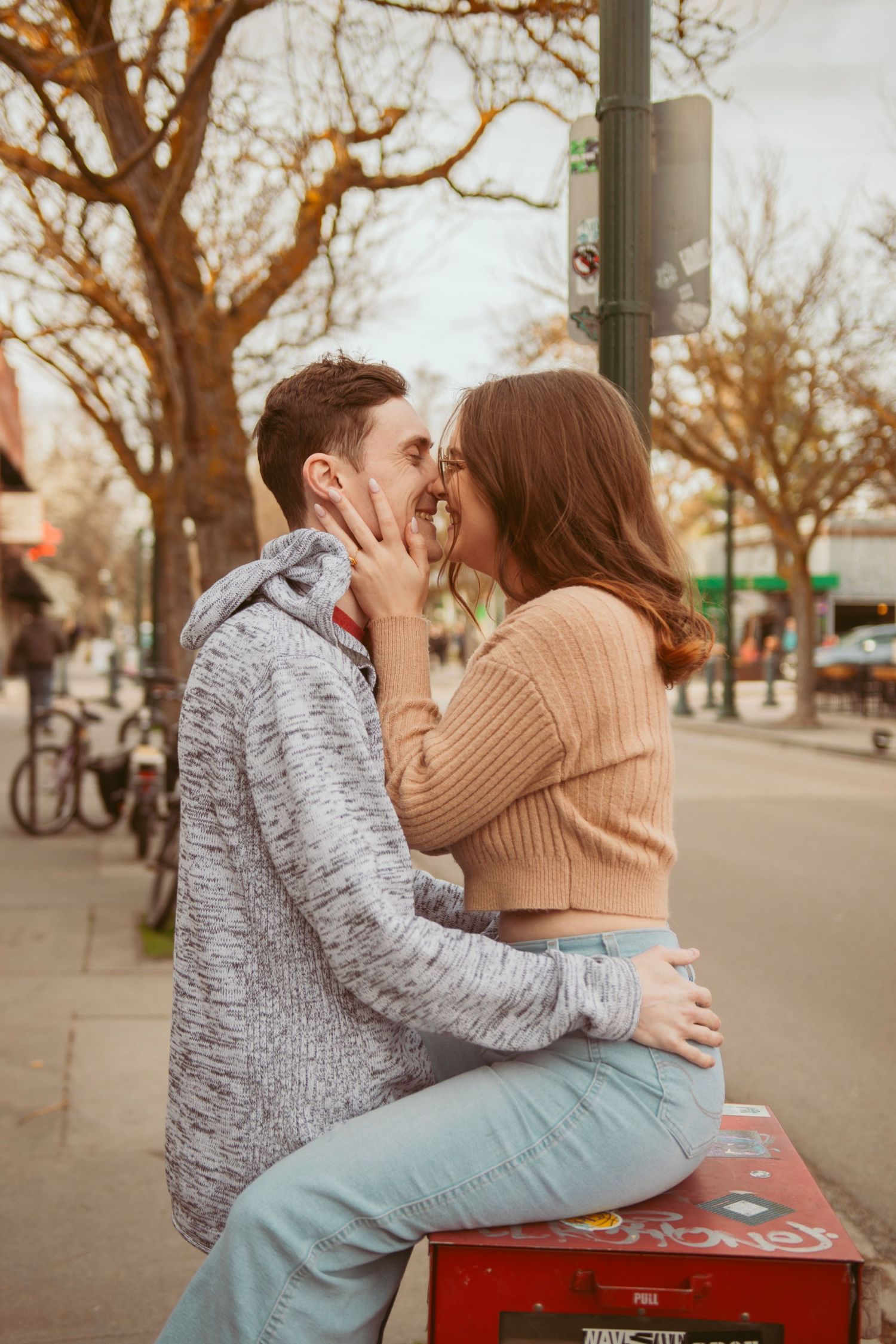 A tender moment between a couple on a red newspaper dispenser on an urban sidewalk.