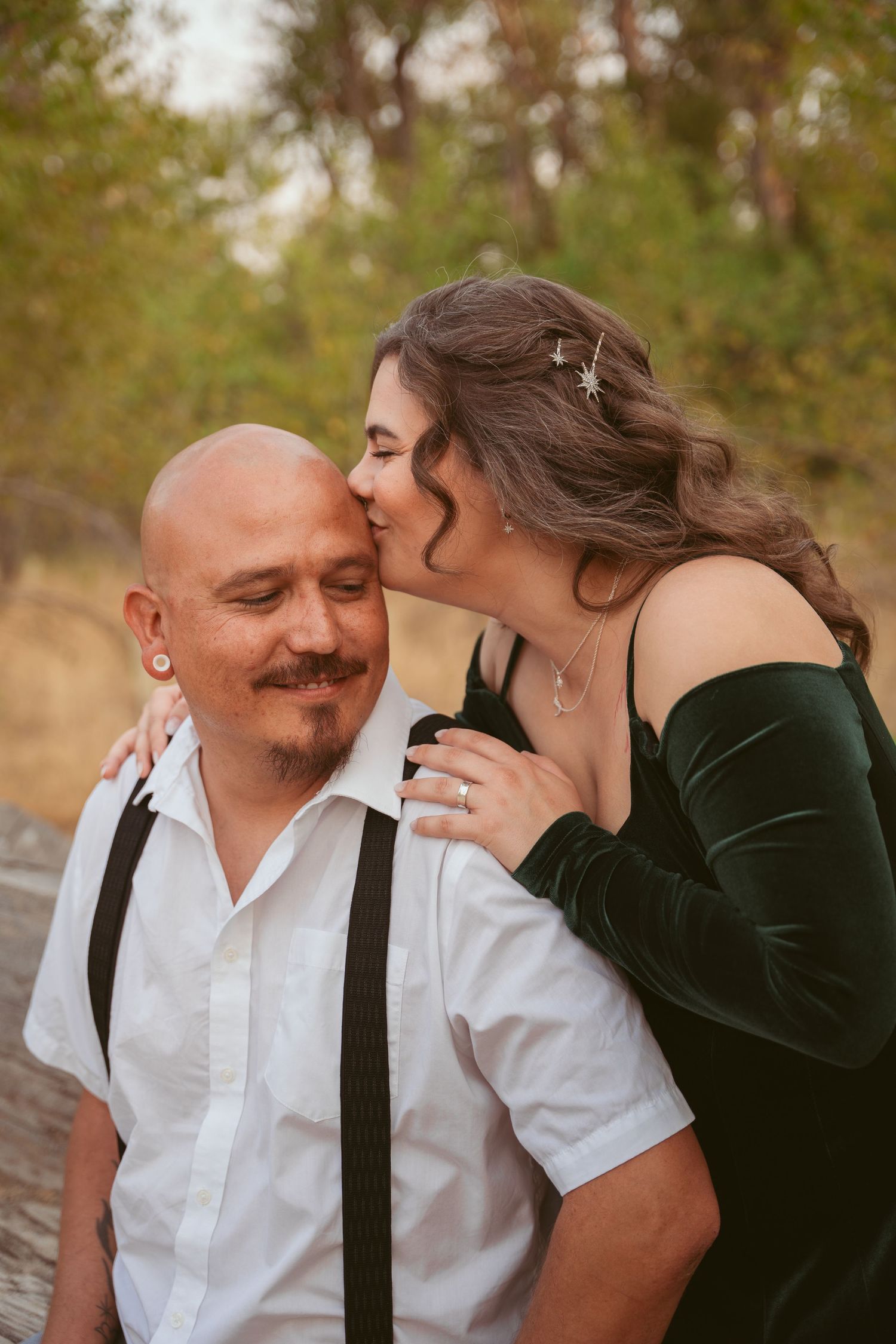 Two people share tender moments during an outdoor photoshoot near trees and natural scenery.