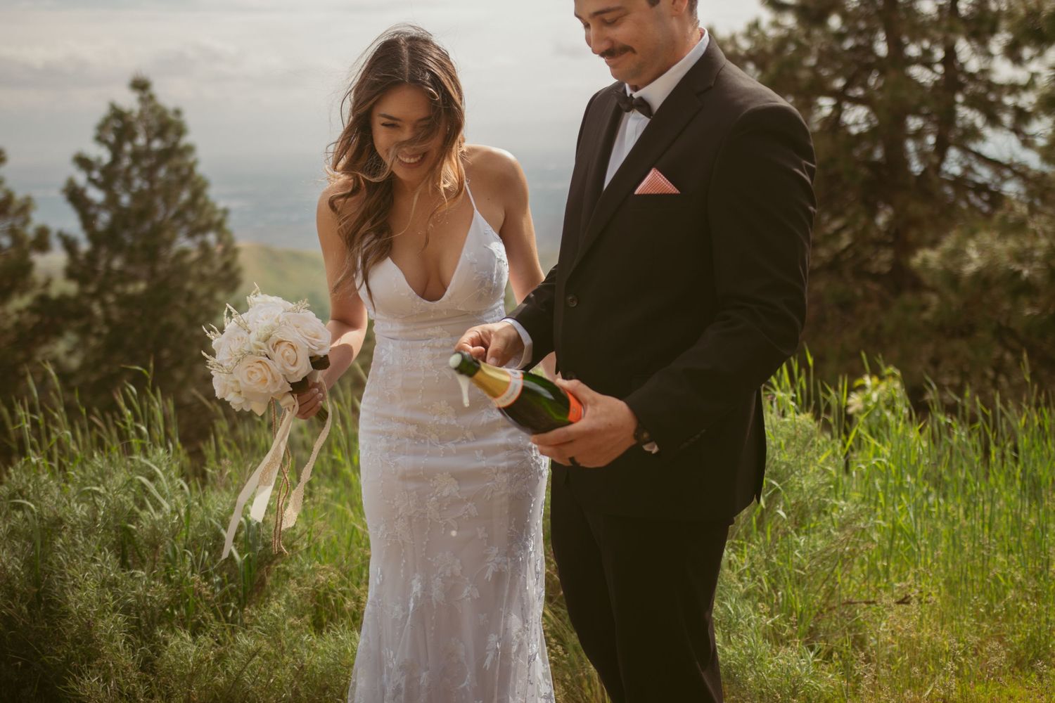 A couple celebrates their wedding day on a scenic mountaintop overlook with champagne and flowers at sunset.