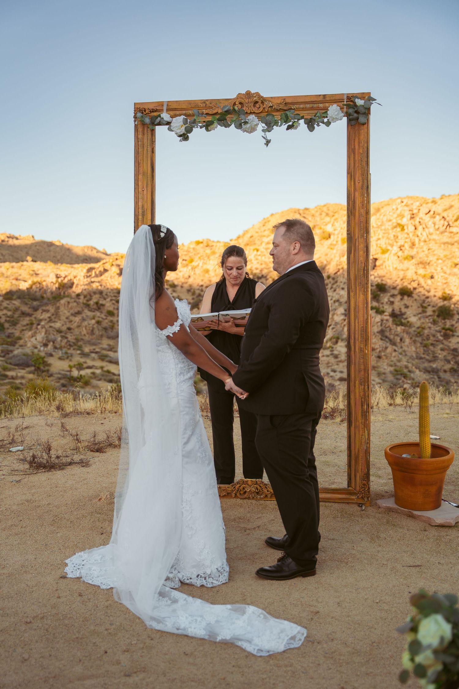 Wedding ceremony under decorated wooden arch frame with mountains and desert landscape behind.