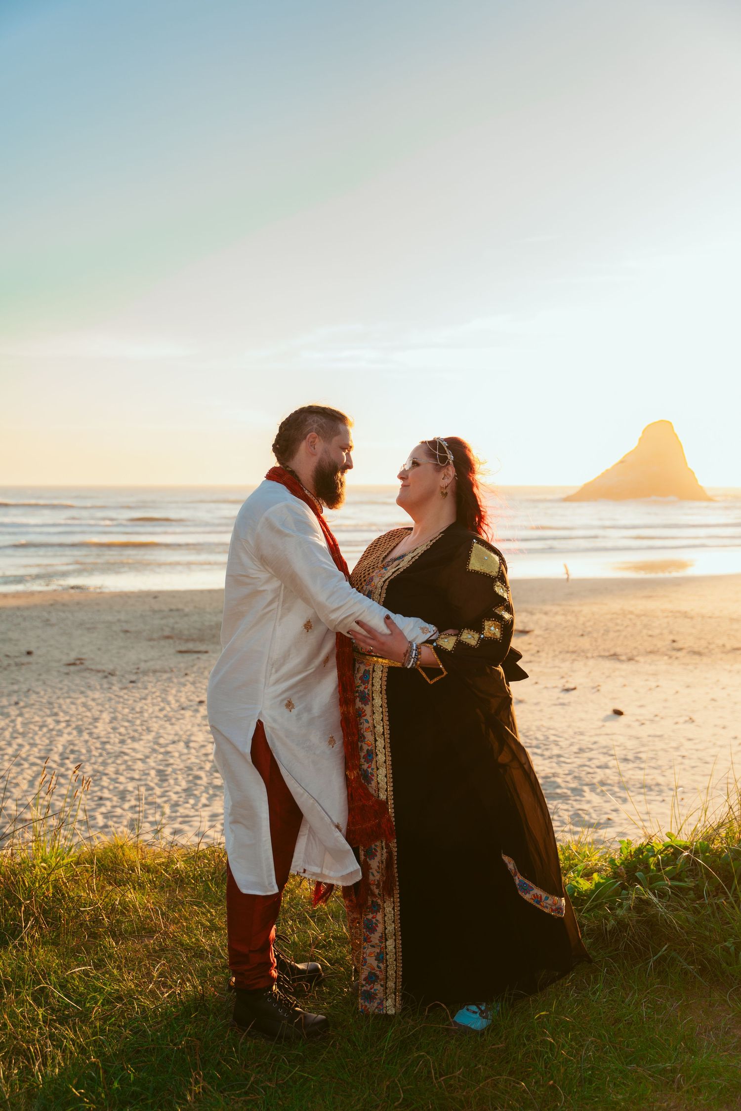 A couple shares an intimate moment on a beach at sunset with coastal rock formations in the distance.