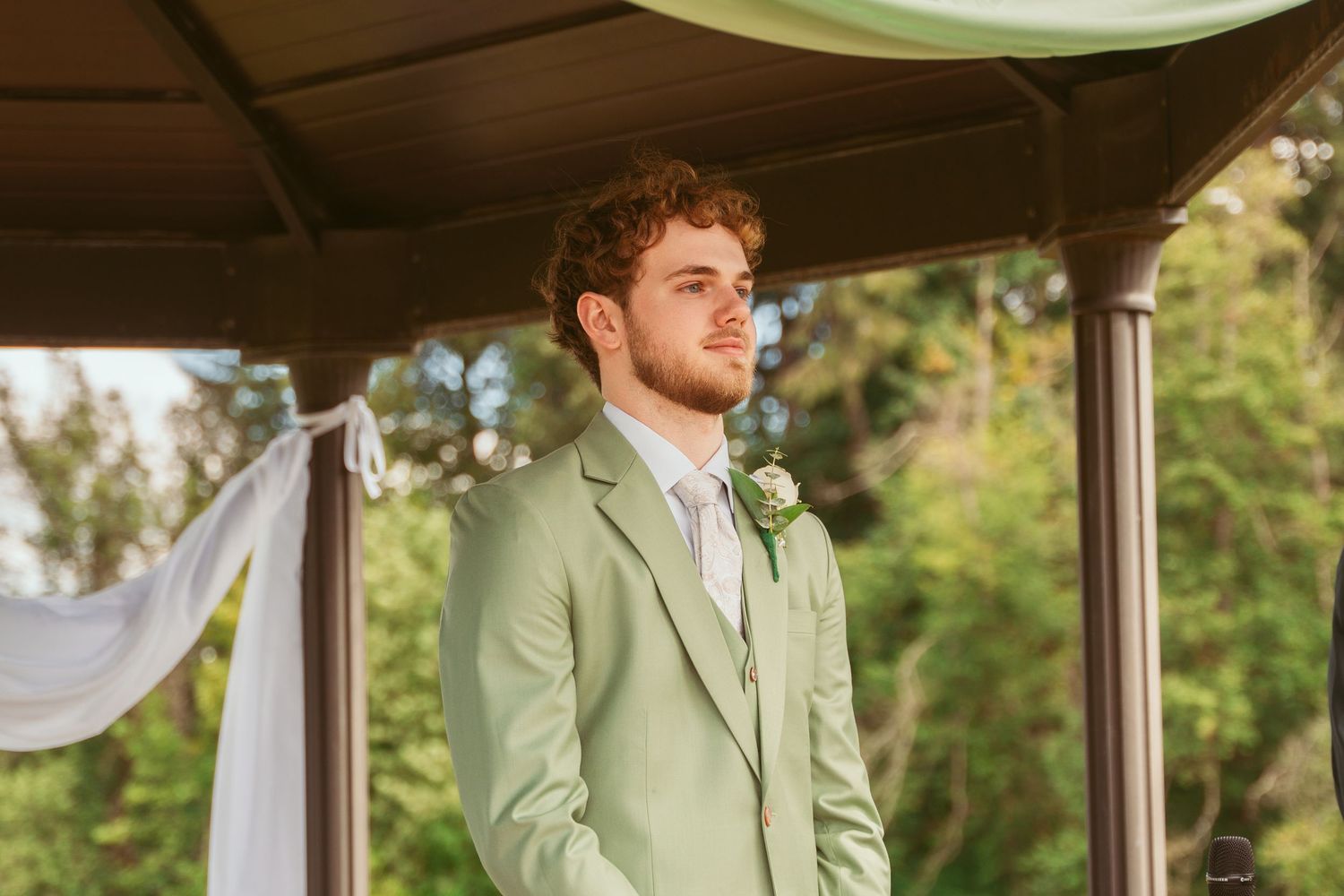 Groom in a mint green suit stands at the altar under a wooden pavilion during an outdoor ceremony.