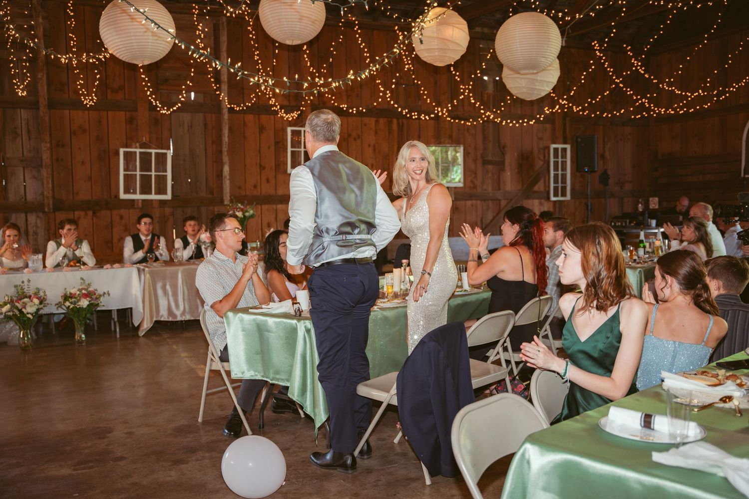 Wedding reception in a rustic barn venue with string lights, paper lanterns, and guests at mint green tables.