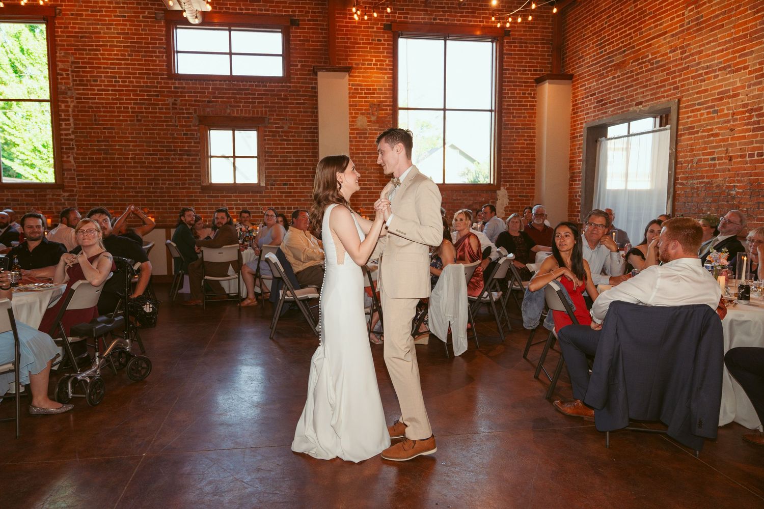 Couple performs first dance in rustic brick venue with string lights and large windows.