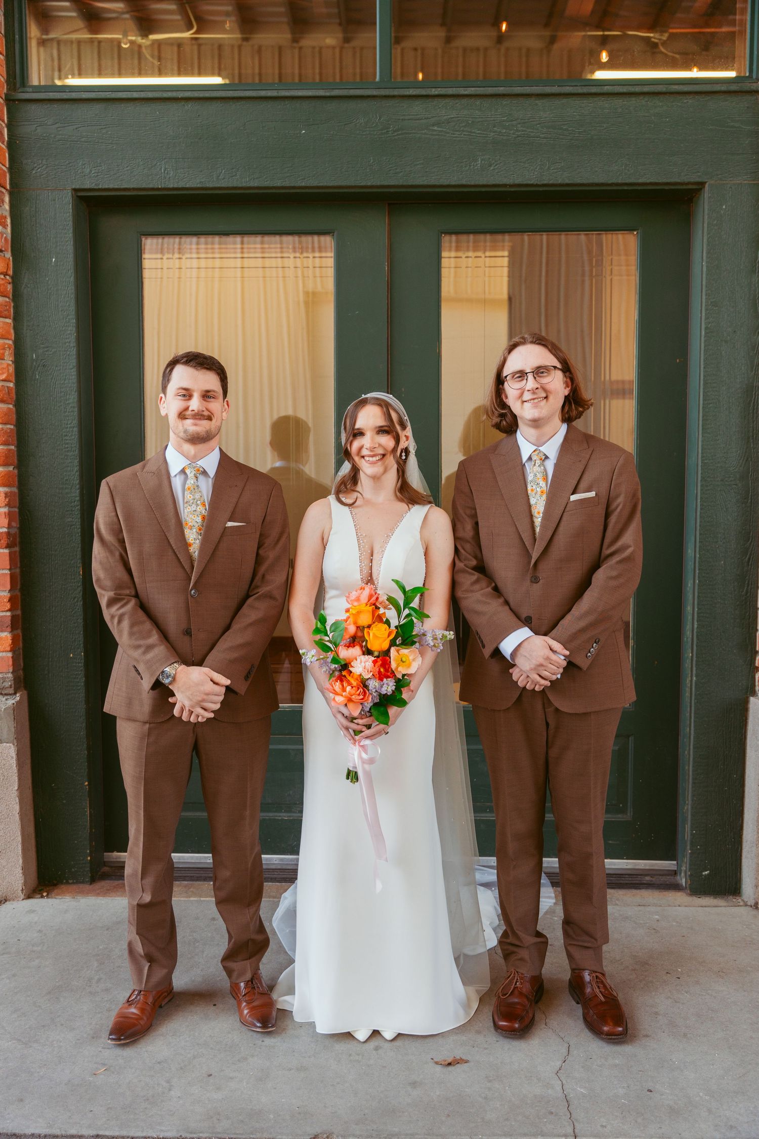 Wedding party lines up against brick building with groomsmen in brown suits and bride holding orange bouquet.