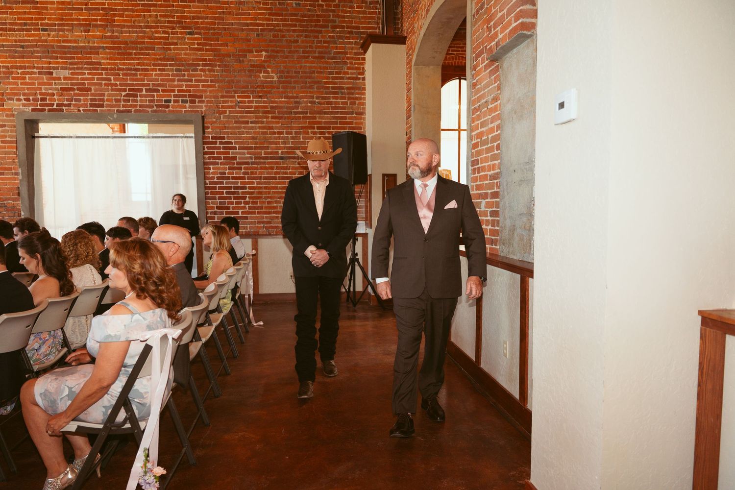 Wedding processional with suited attendants walking down aisle at brick-walled venue.