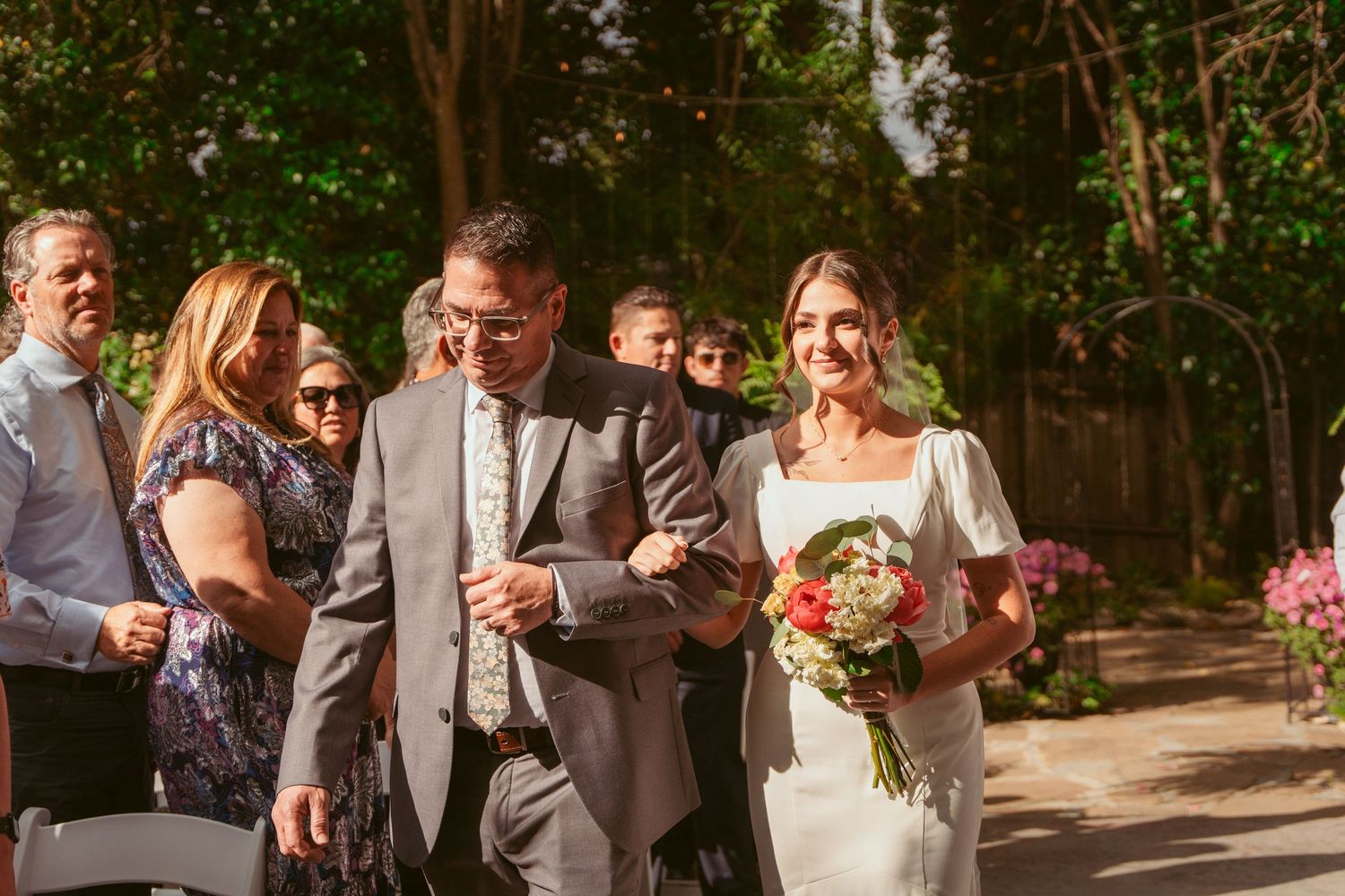 Wedding procession walking through garden pathway with pink floral bouquets.