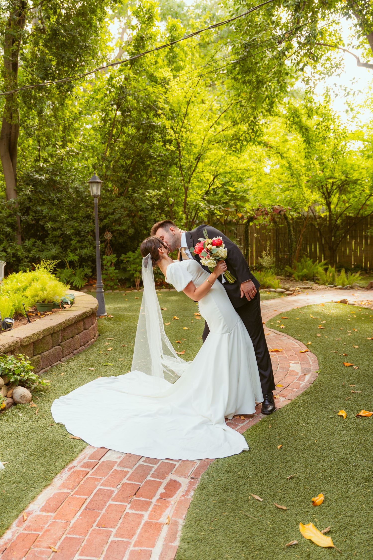A romantic wedding moment captured in a lush garden setting with a flowing white dress and brick pathway.