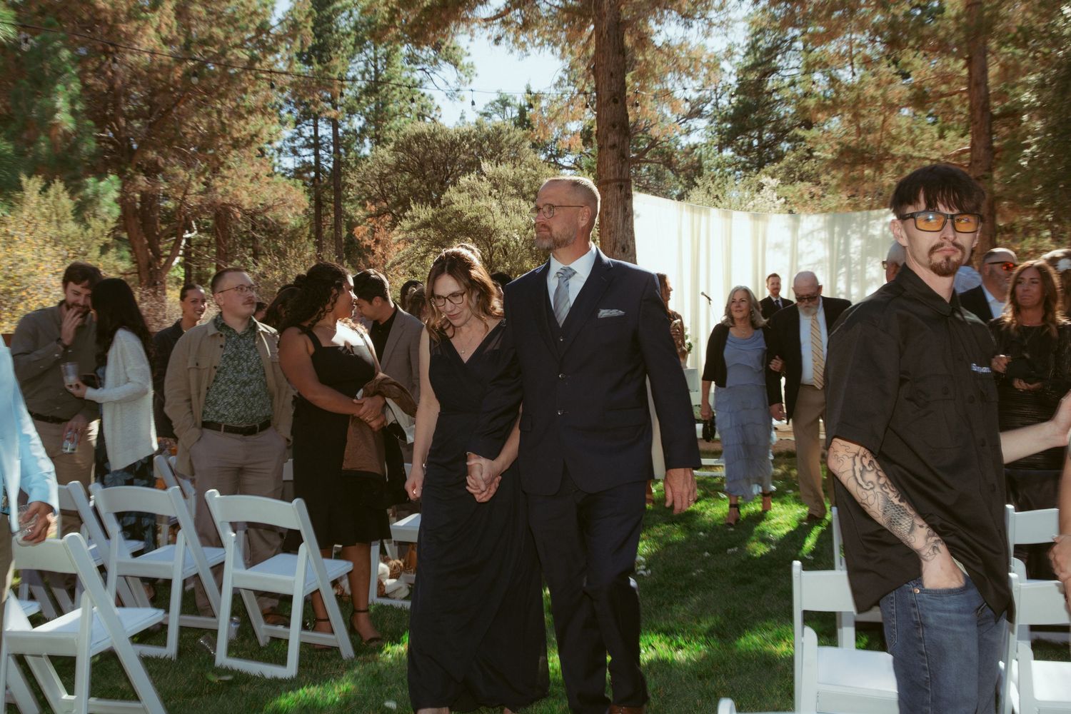 Wedding guests mingle outdoors during a ceremony setup with white chairs under tall pine trees.