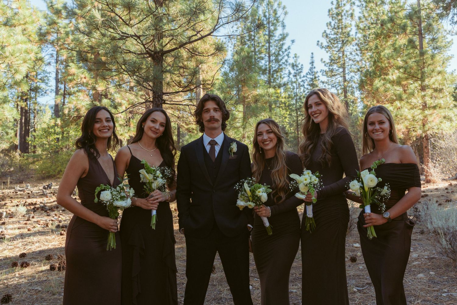 Bridal party poses together in outdoor forest setting with pine trees in background.