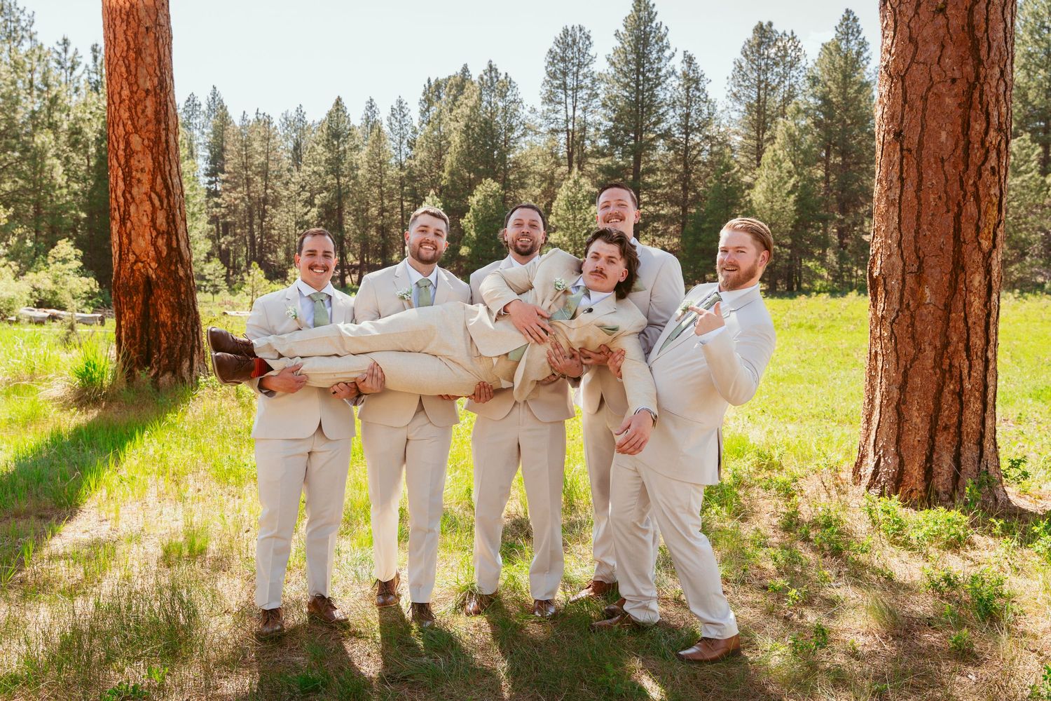 Wedding party members in matching cream suits pose playfully in an outdoor forest setting.