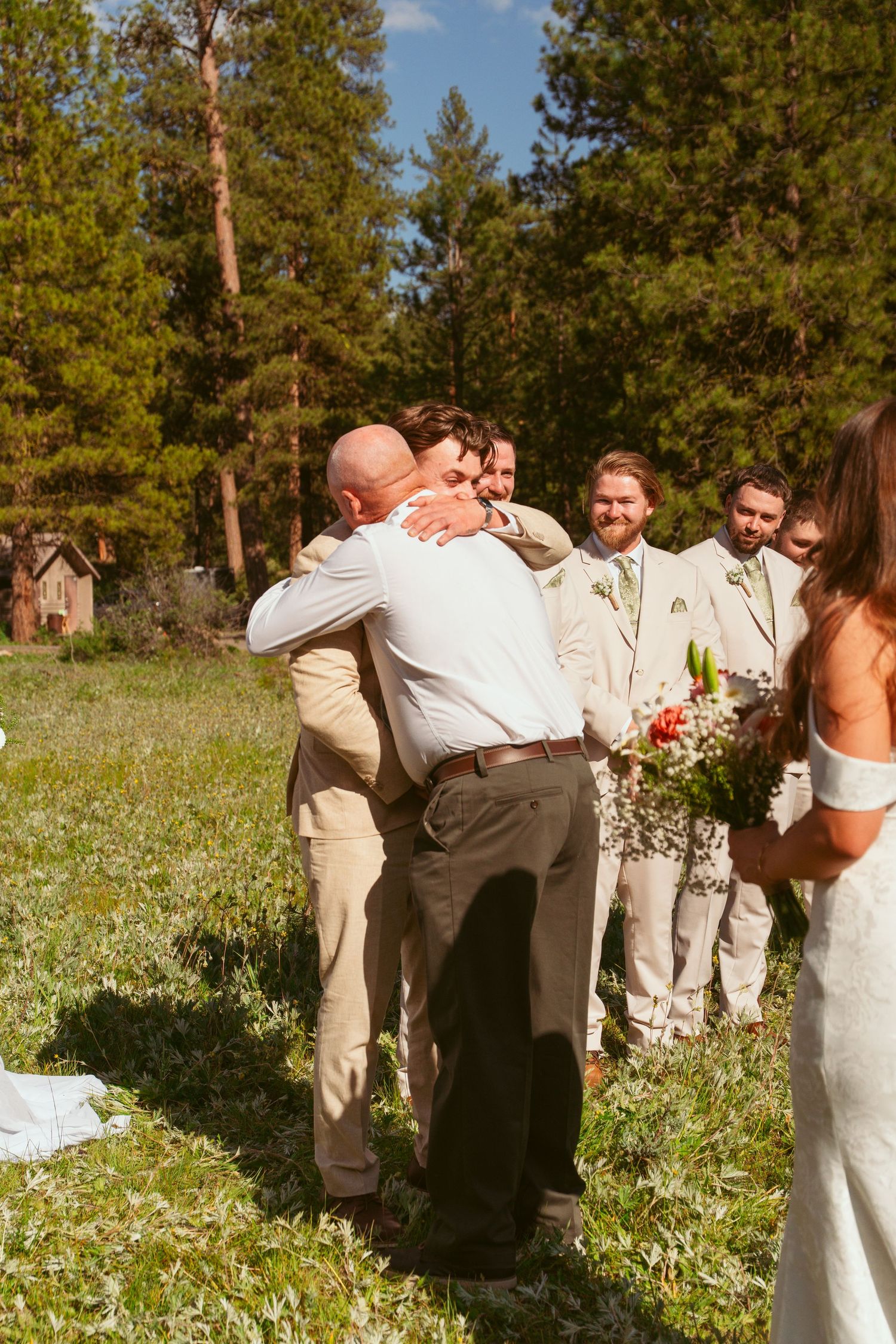 Wedding party celebrates in a sunny mountain meadow surrounded by tall pine trees.