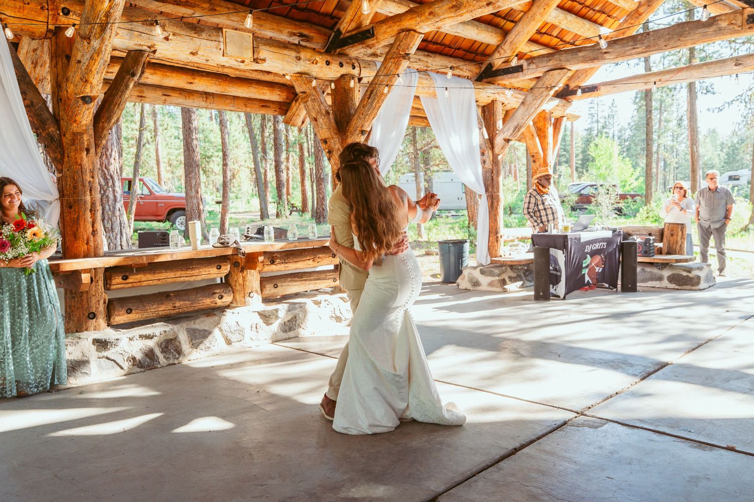 Rustic outdoor wedding venue with exposed wooden beams and flowing white curtains in forest setting.