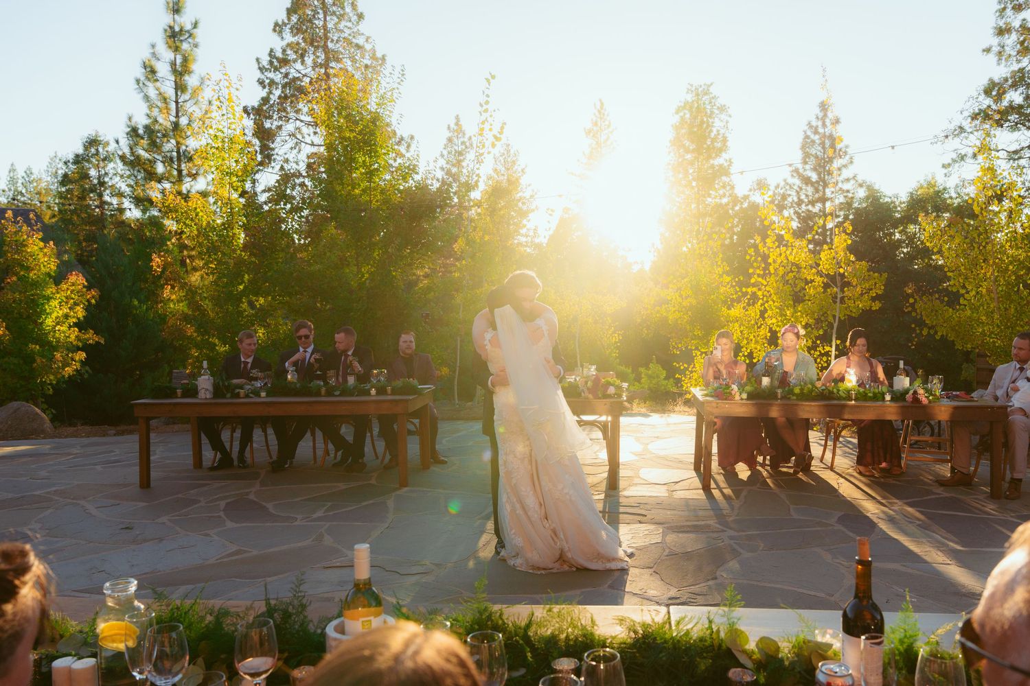 Couple dancing at sunset on an outdoor stone patio surrounded by tall pine trees and wooden benches.