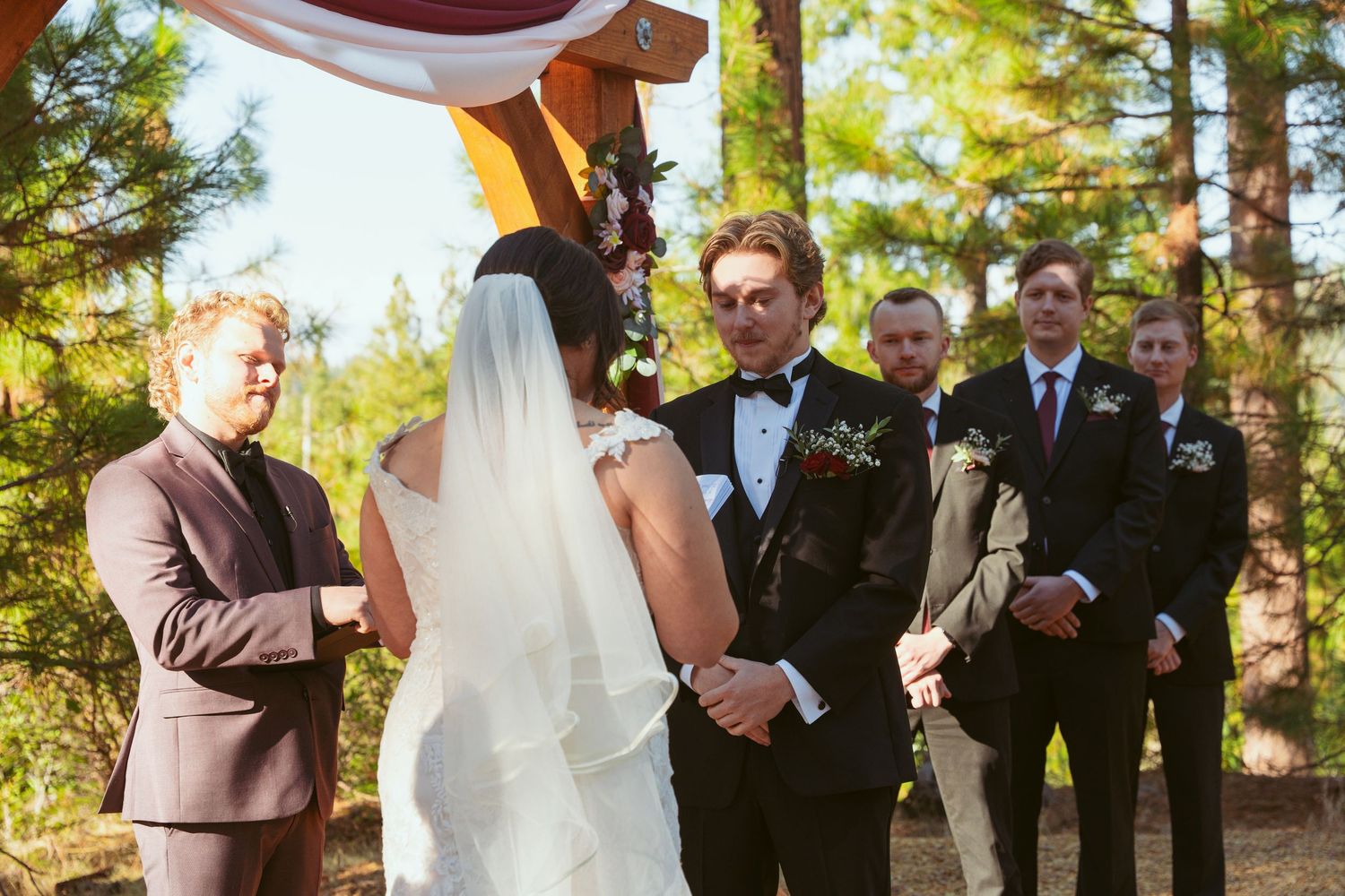 Wedding ceremony with groomsmen and officiant standing beneath pine trees.